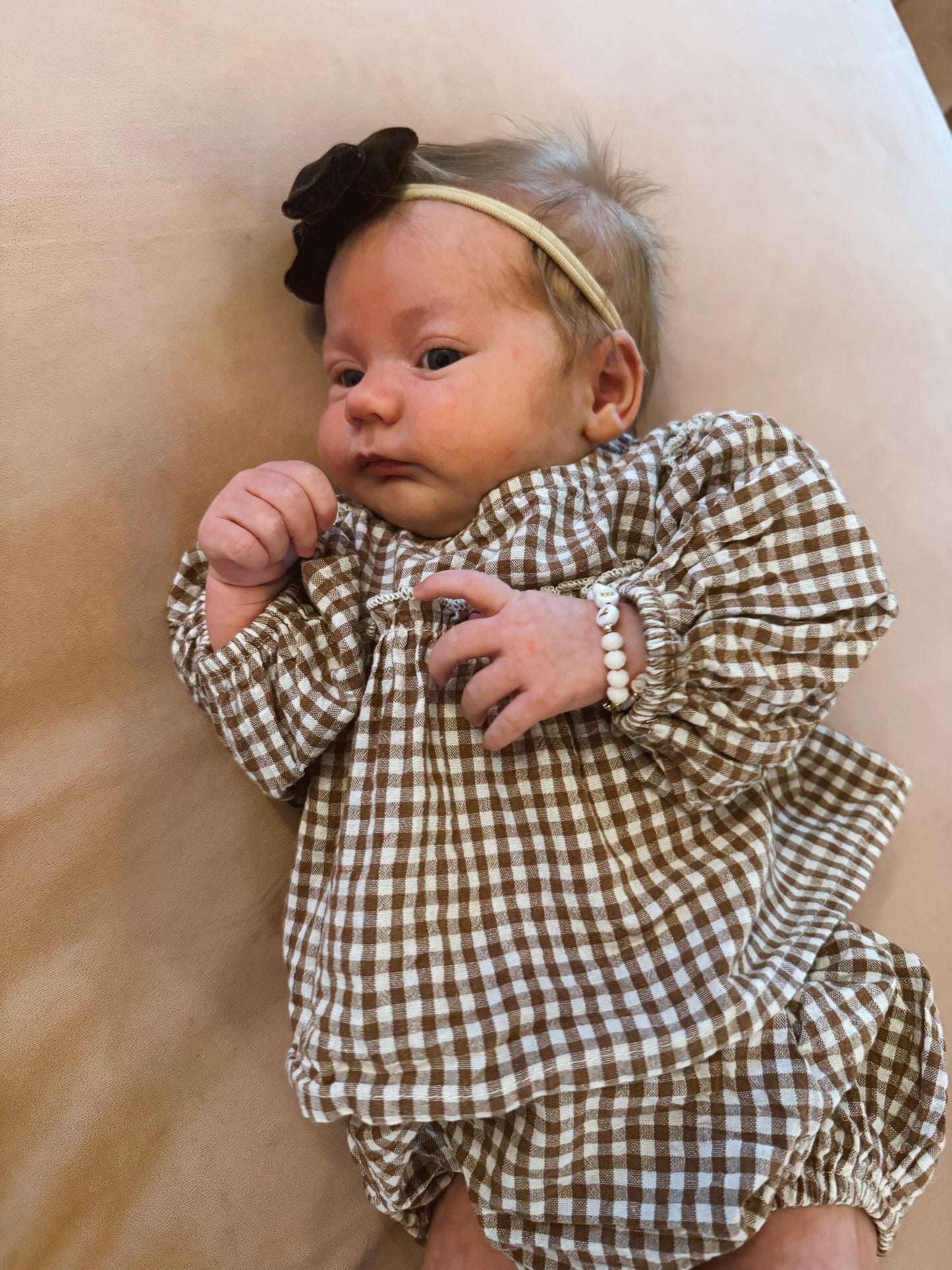A baby in the forever french baby Bloomer Set | Honey Gingham, with a brown headband featuring a flower and a white beaded bracelet, lies on a light surface looking slightly to the side.
