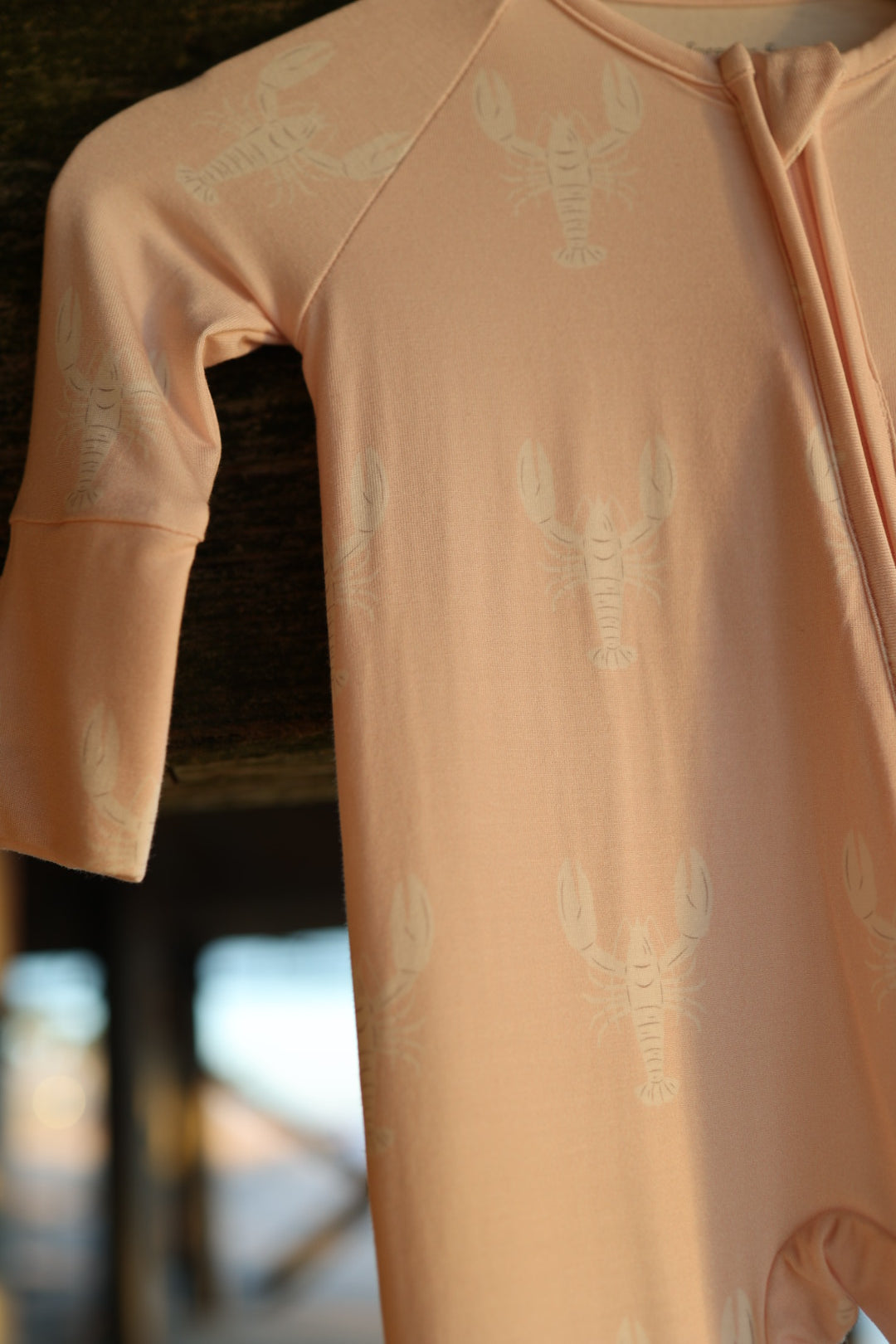 The forever french baby Bamboo Zip Pajamas | Lobster in peach, featuring a subtle white lobster print and a visible sleeve with front zipper, hangs against a softly blurred background.