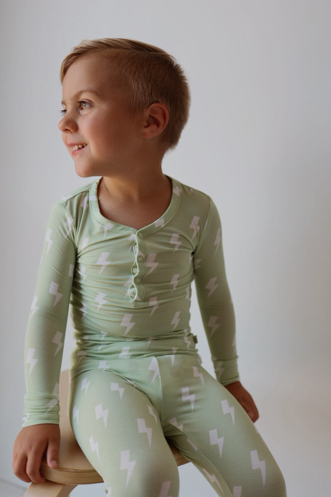 A young child with short light brown hair, smiling and sitting on a wooden stool, wears forever french baby’s Bamboo Two Piece Pajamas | Flash—light green pajamas with white lightning bolts—against a plain background.