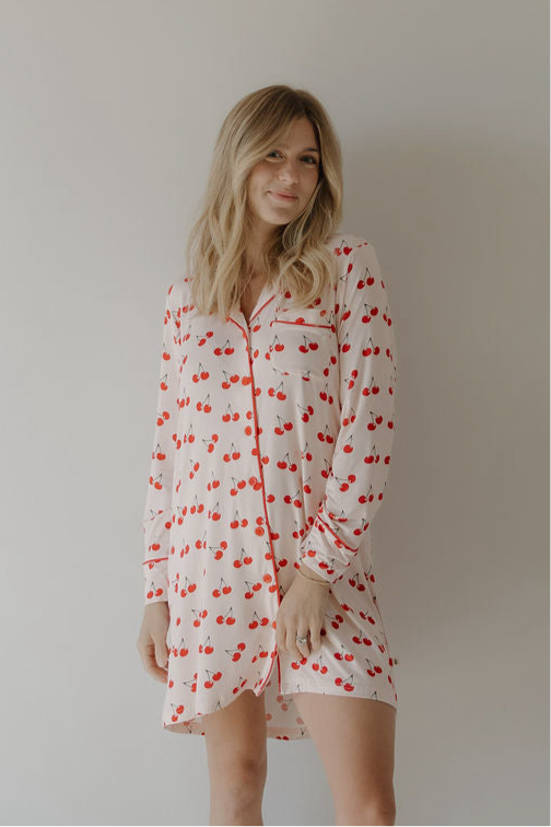 A woman with blonde hair smiles softly in a forever french baby Women's Bamboo Sleeping Dress | Cherry Orchard, featuring long sleeves and red cherry patterns, standing against a plain light-colored wall.