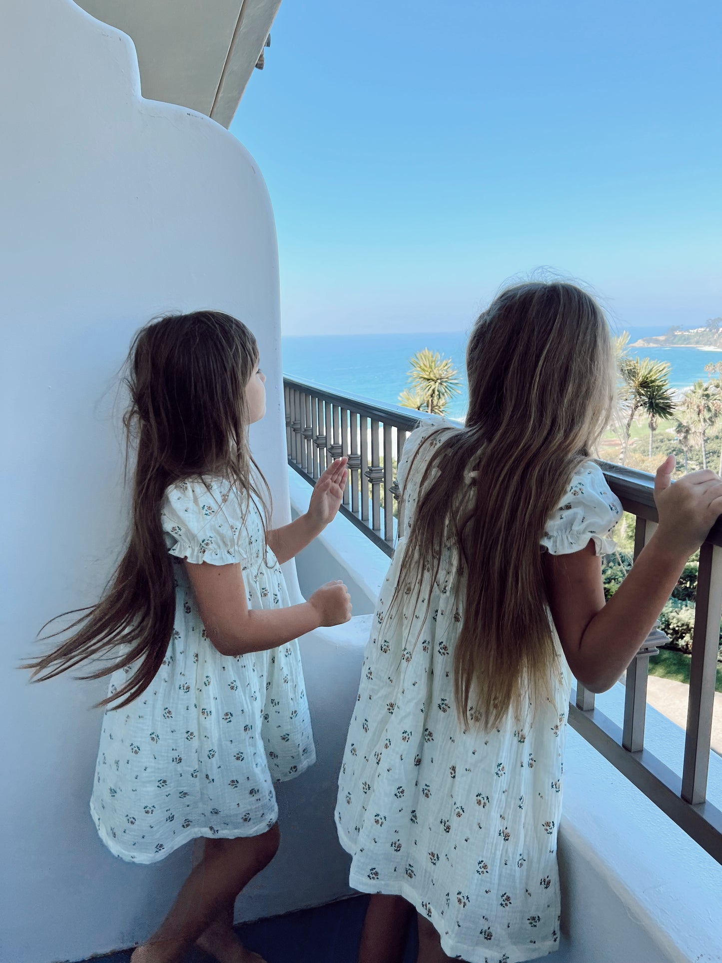 Two young girls with long hair, dressed in matching "Garden Days" ivory muslin dresses adorned with floral prints by forever french baby, stand on a balcony overlooking a serene blue ocean and sandy coastline. They gaze out towards the view, with their backs and profiles partially visible in the clear, sunny weather.