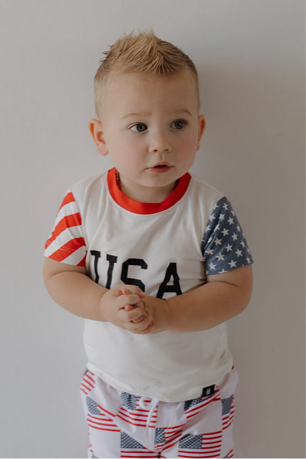A young boy with short blond hair wears the forever french baby Child Bamboo T-Shirt | Stars & Stripes, featuring a breathable USA-inspired design, as he stands against a white background looking to the side with hands clasped.