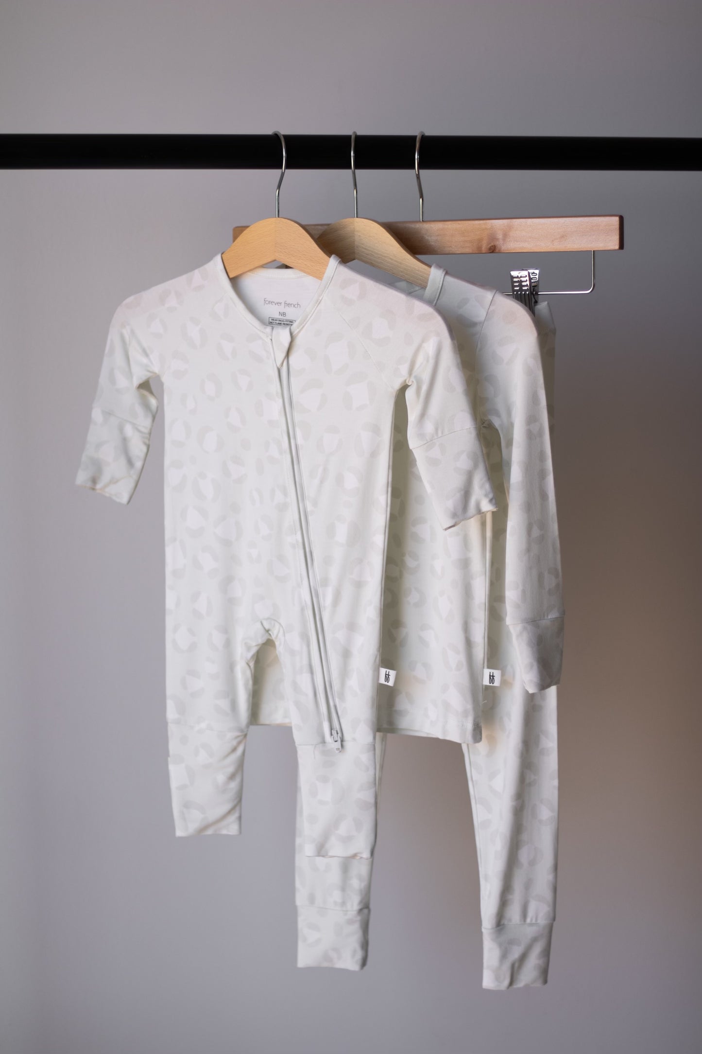 Two light-colored Bamboo Zip Pajamas | Meow by forever french baby, featuring subtle patterns and long sleeves, hang on wooden hangers from a black rack against a gray background, highlighting breathable sleepwear for babies.