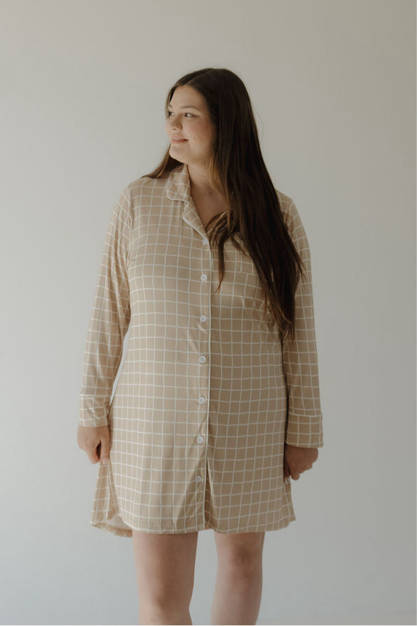 A woman with long brown hair wears the forever french baby Women's Bamboo Sleeping Dress | Grid, a beige, long-sleeved, button-down nightdress with a white checkered pattern. She stands against a plain light background, smiling softly.