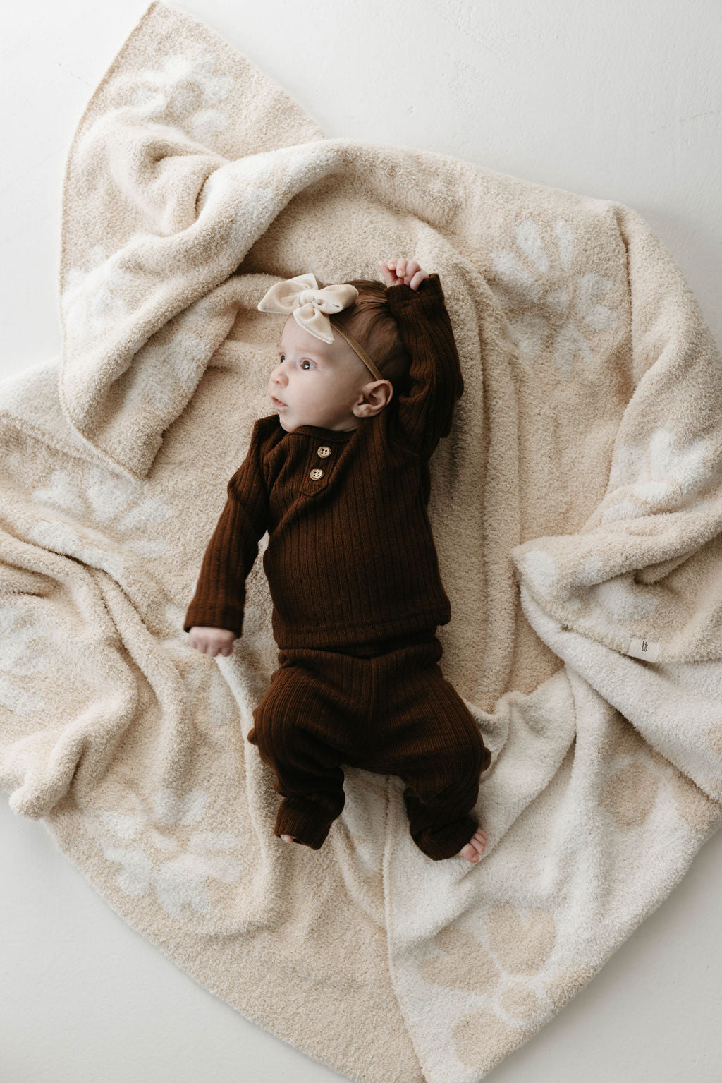 A baby dressed in a brown ribbed onesie and matching pants lies on the Plush Blanket | Lazy Daisy by forever french baby, a beige blanket adorned with a soft floral pattern, crafted from velvety-rich microfiber feather yarn. The baby wears a cream-colored headband with a bow and looks to the side with one arm extended above their head. The background is neutral-toned.