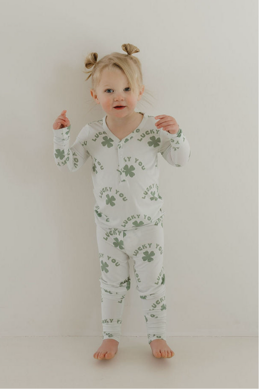 A young child with light hair styled in two buns stands barefoot against a white wall, wearing forever french baby’s Bamboo Two Piece Pajamas | Lucky You—soft, breathable green shamrock print for cozy sleep.