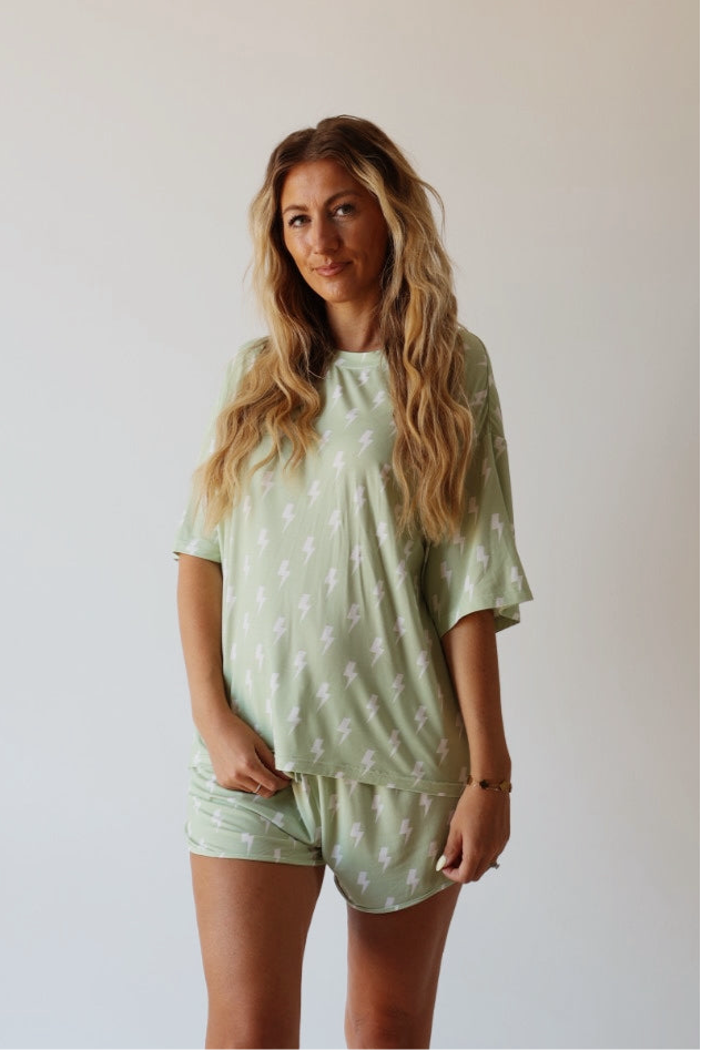 A woman with long, wavy blonde hair wears the Women's Bamboo Short Set | Flash by forever french baby, featuring a light green fabric with white lightning bolts. She stands against a plain light background, smiling softly at the camera.