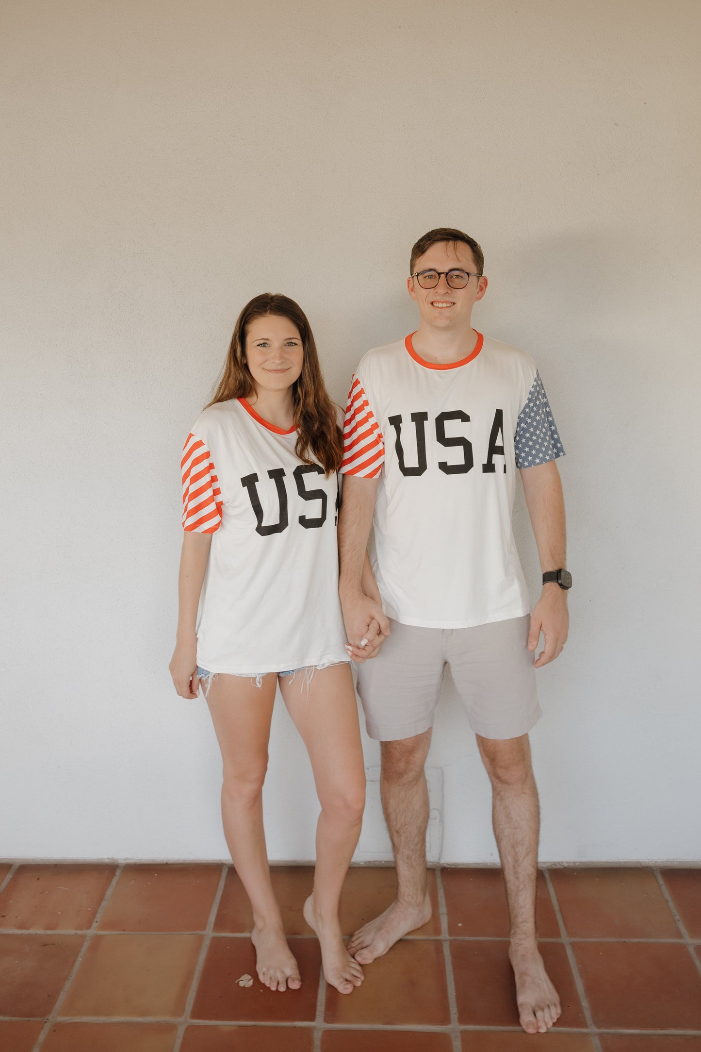 A woman and man stand barefoot on a tiled floor, holding hands, both wearing matching forever french baby Adult Bamboo T-Shirts | Stars & Stripes with red, white, and blue details.