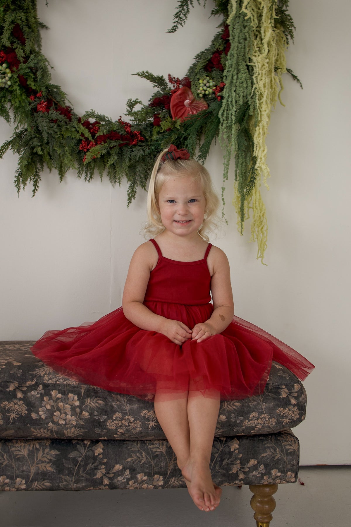 A young girl wearing a cranberry red ff Tutu Dress | Cranberry with a ribbed top and a layered tulle skirt sits on a patterned bench in front of a holiday wreath.