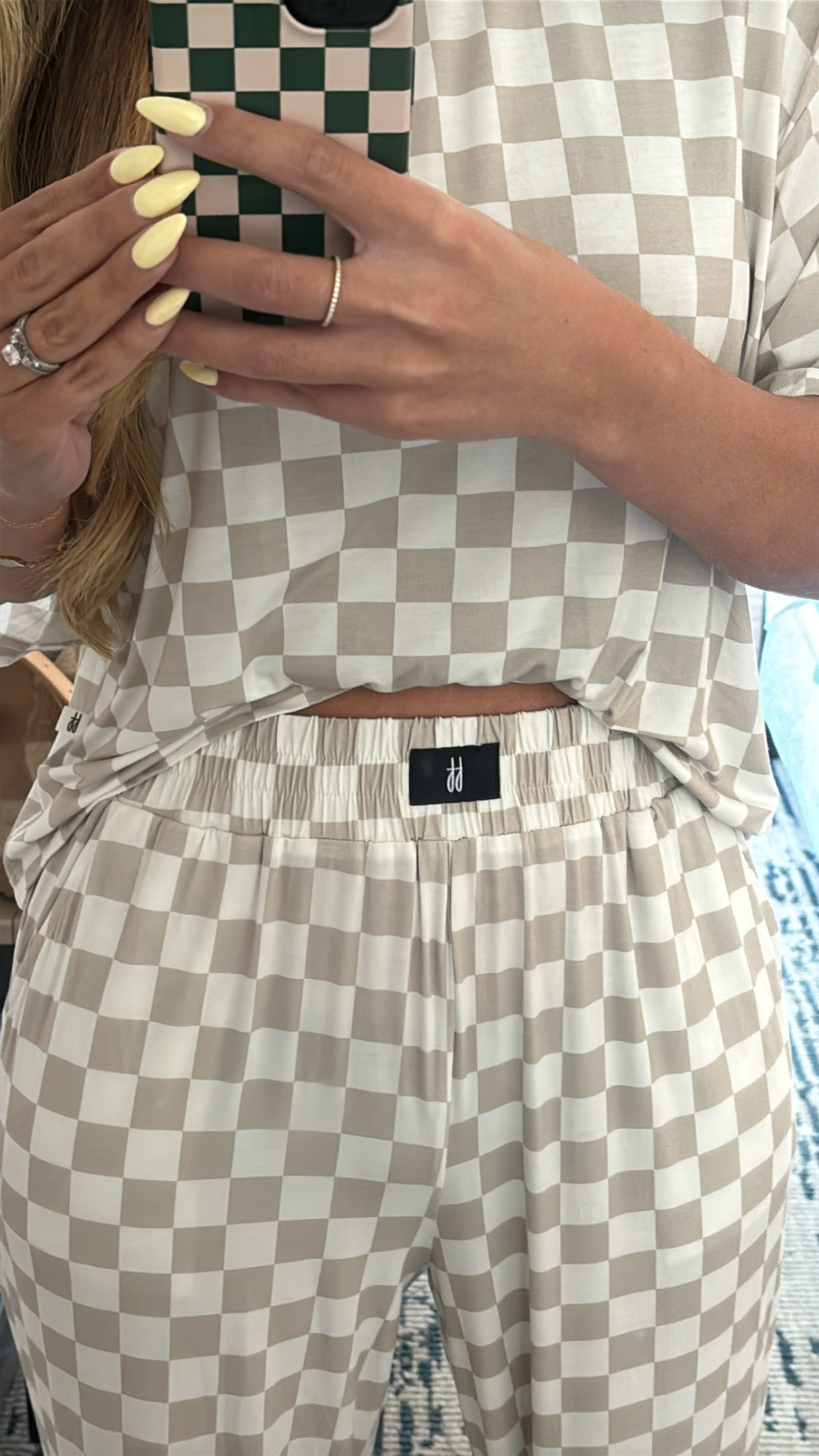 A person wearing the forever french baby Women's Boxy Tee Lounge Set in beige and white checkerboard takes a mirror selfie, highlighting their yellow manicured nails and a ring on their left hand.