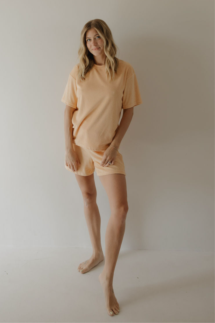 A woman with wavy blonde hair stands barefoot by a plain wall, wearing the Women's Terry Short Set in Sherbert from forever french baby, her peach outfit radiating effortless comfort as she smiles gently.