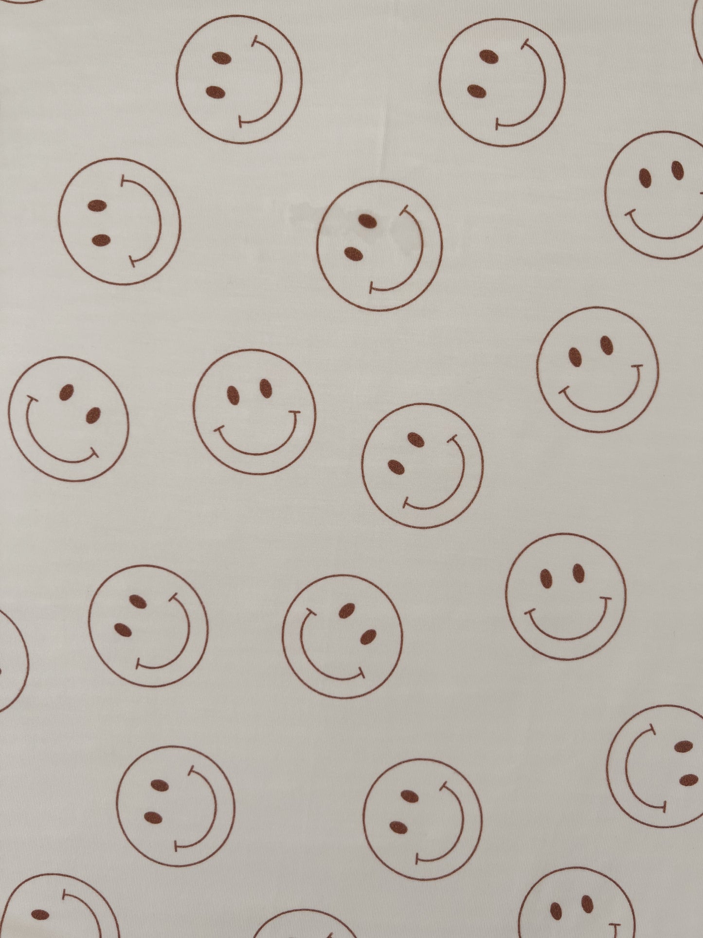 The Bamboo Swaddle | Just Smile from forever French baby is a white, hypo-allergenic fabric adorned with a pattern of evenly spaced brown smiley faces. Each smiley face features two eyes and a curved mouth within a circle, ensuring consistent size and appearance. Ideal for year-round comfort, this swaddle is perfect for your little one.