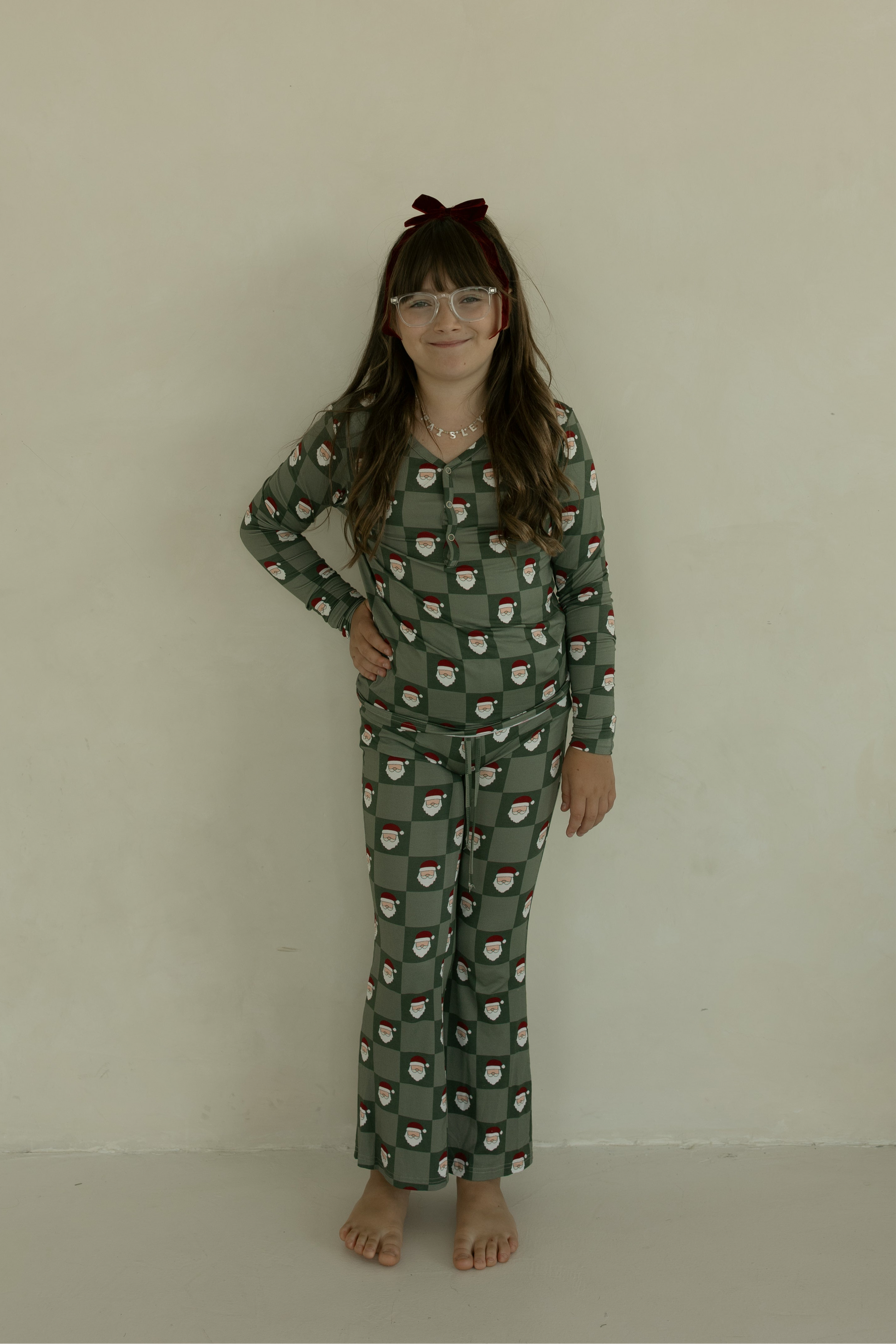 A young girl with long brown hair, a red bow headband, and glasses smiles at the camera in forever french baby Pre-Teen Flare Bamboo Pajamas | Santa Checkerboard, standing barefoot against a plain light wall.