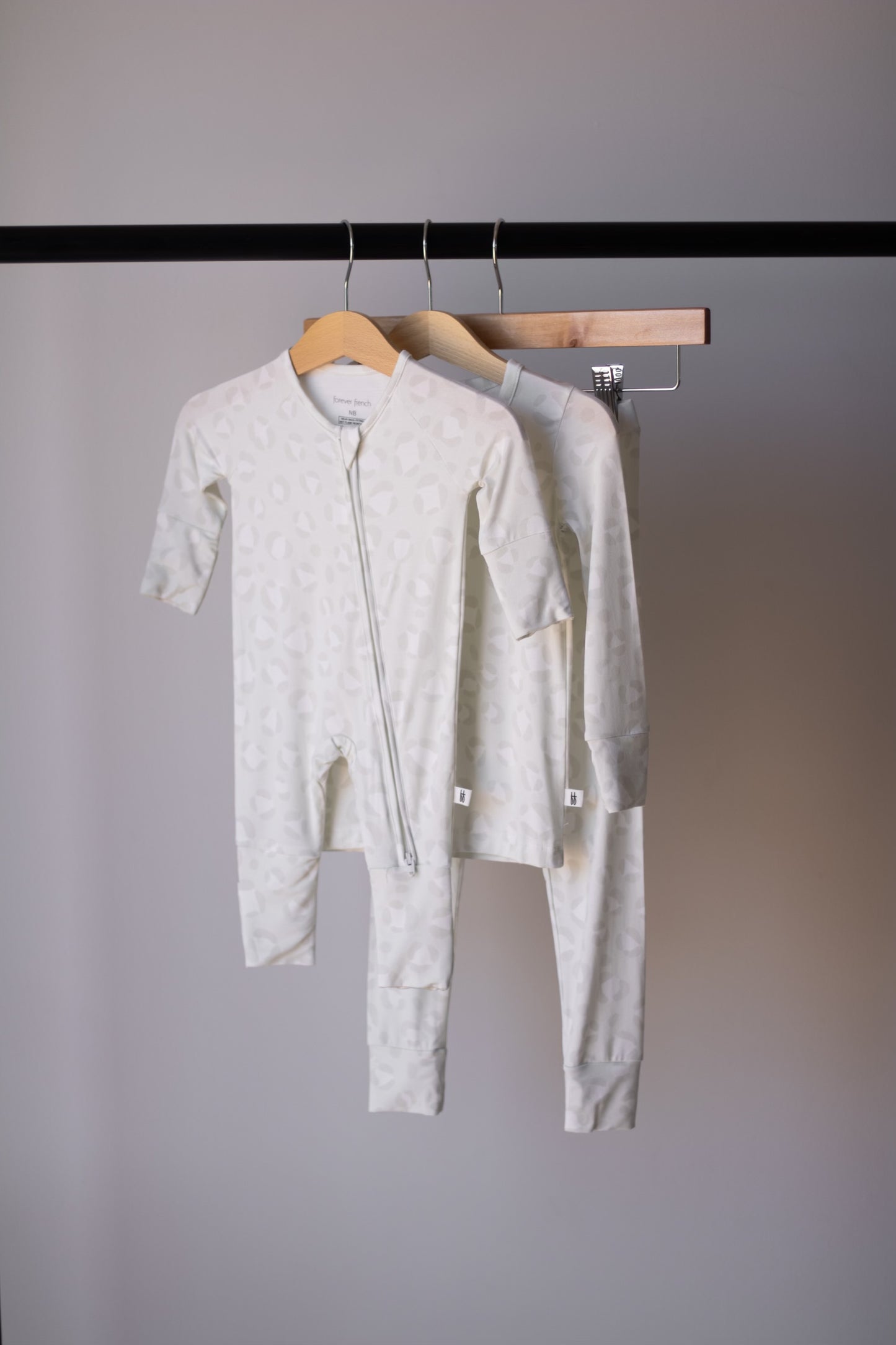 Two light-colored Bamboo Two Piece Pajamas | Meow by forever french baby, featuring subtle patterns and long sleeves for babies and toddlers, hang on wooden hangers from a black clothing rack against a plain gray background.