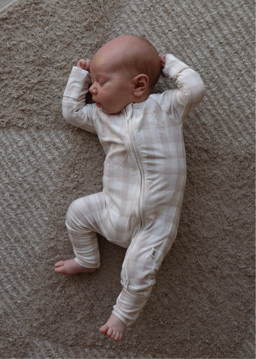 A newborn, dressed in forever french baby Bamboo Zip Pajamas | Gingham, sleeps with arms raised and legs relaxed on a textured beige carpet.