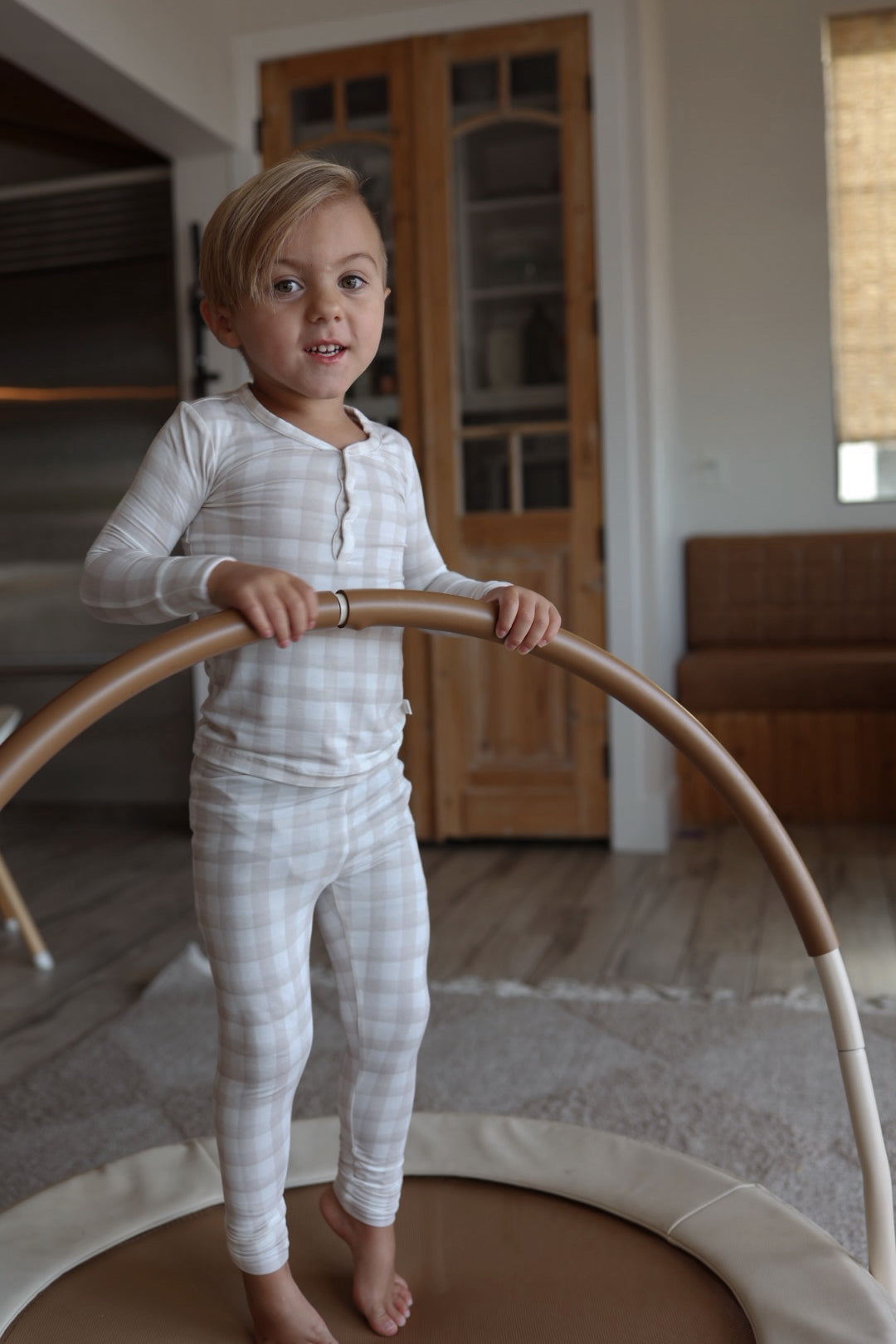 A young child with blond hair, barefoot on a round play mat, wears forever french baby’s Bamboo Two Piece Pajamas in Gingham while holding a circular climbing frame indoors.