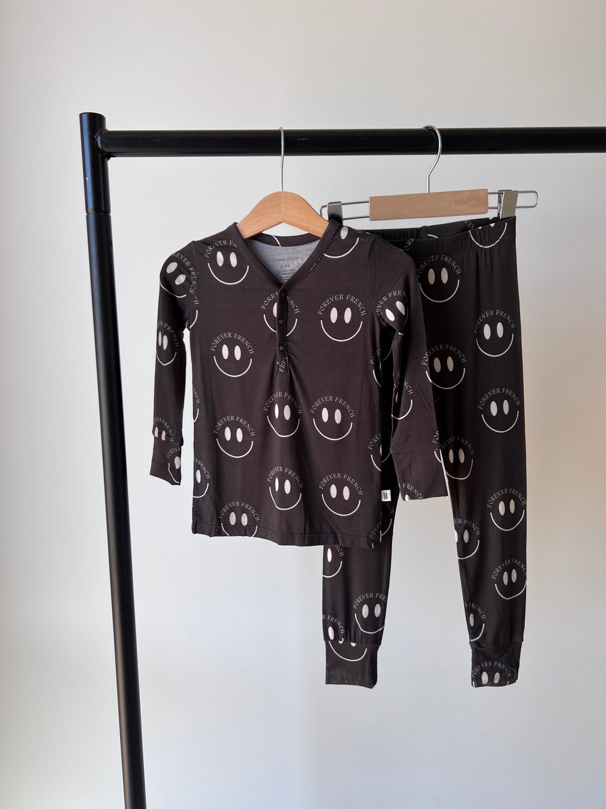 A Bamboo Two Piece Pajama set from forever french baby is hanging on a black horizontal bar. The breathable sleepwear is charcoal and white featuring a pattern of large smiling faces with accompanying circular text. The top has long sleeves and a partial button-front design, while the pants come with an elastic waistband.