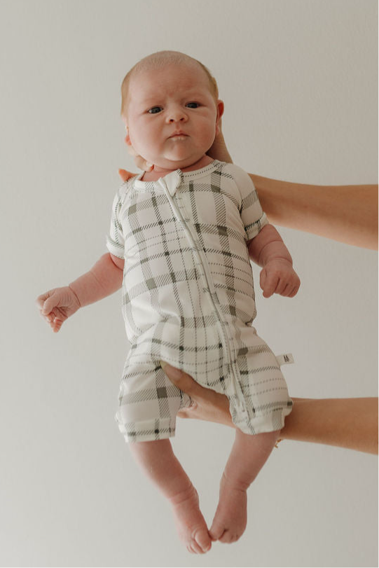 A baby wearing the forever french baby Bamboo Zip Shortie in Picnic Plaid is held up by an adult's hands against a plain light background, looking directly at the camera with a neutral expression.