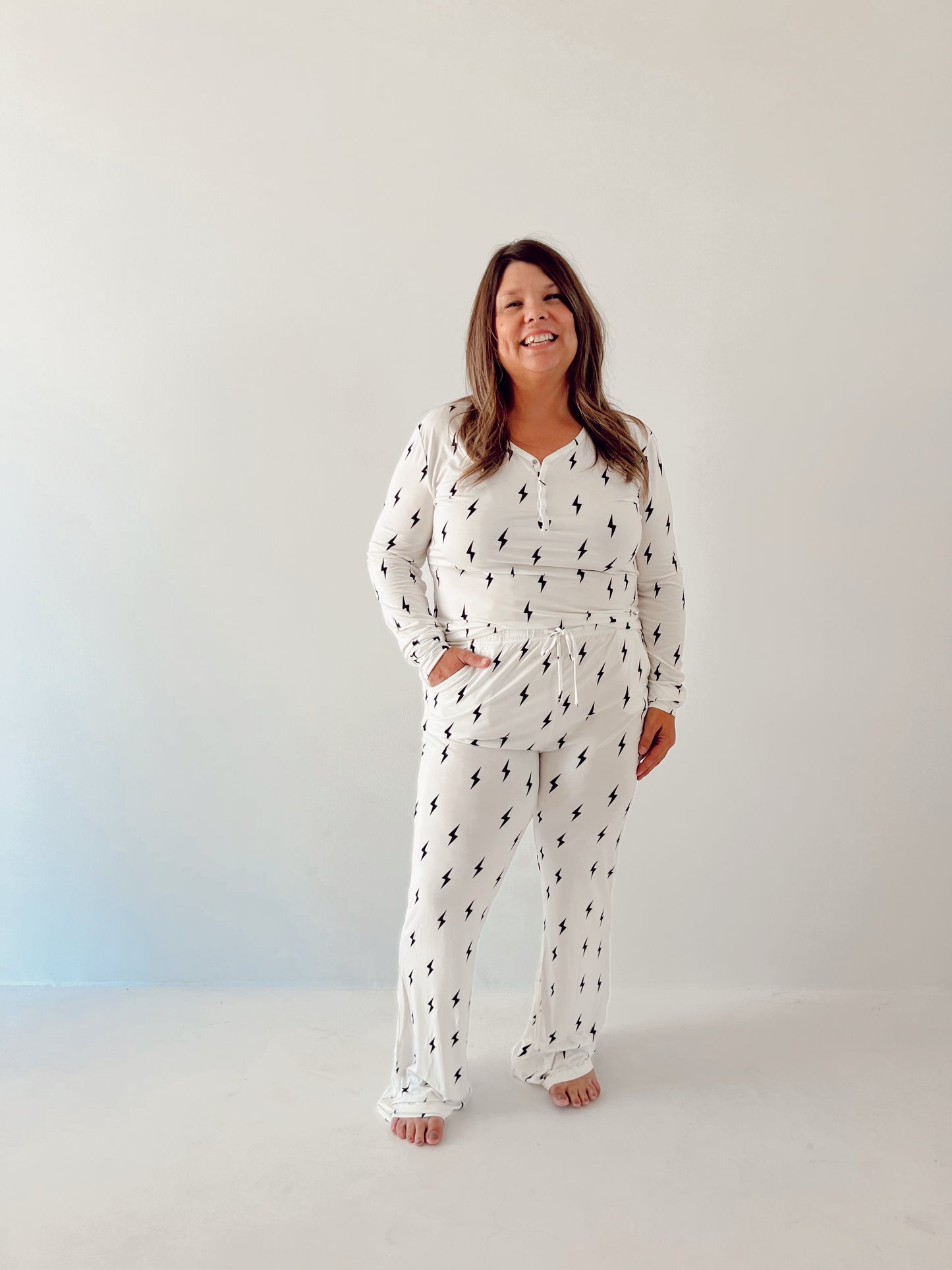 A woman smiles at the camera, dressed in a forever french baby Women's Bamboo Pajamas in white with a black lightning bolt print. She stands barefoot against a plain, light-colored background with her left hand in her pocket. These bamboo pajamas are breastfeeding-friendly, making them both stylish and practical.