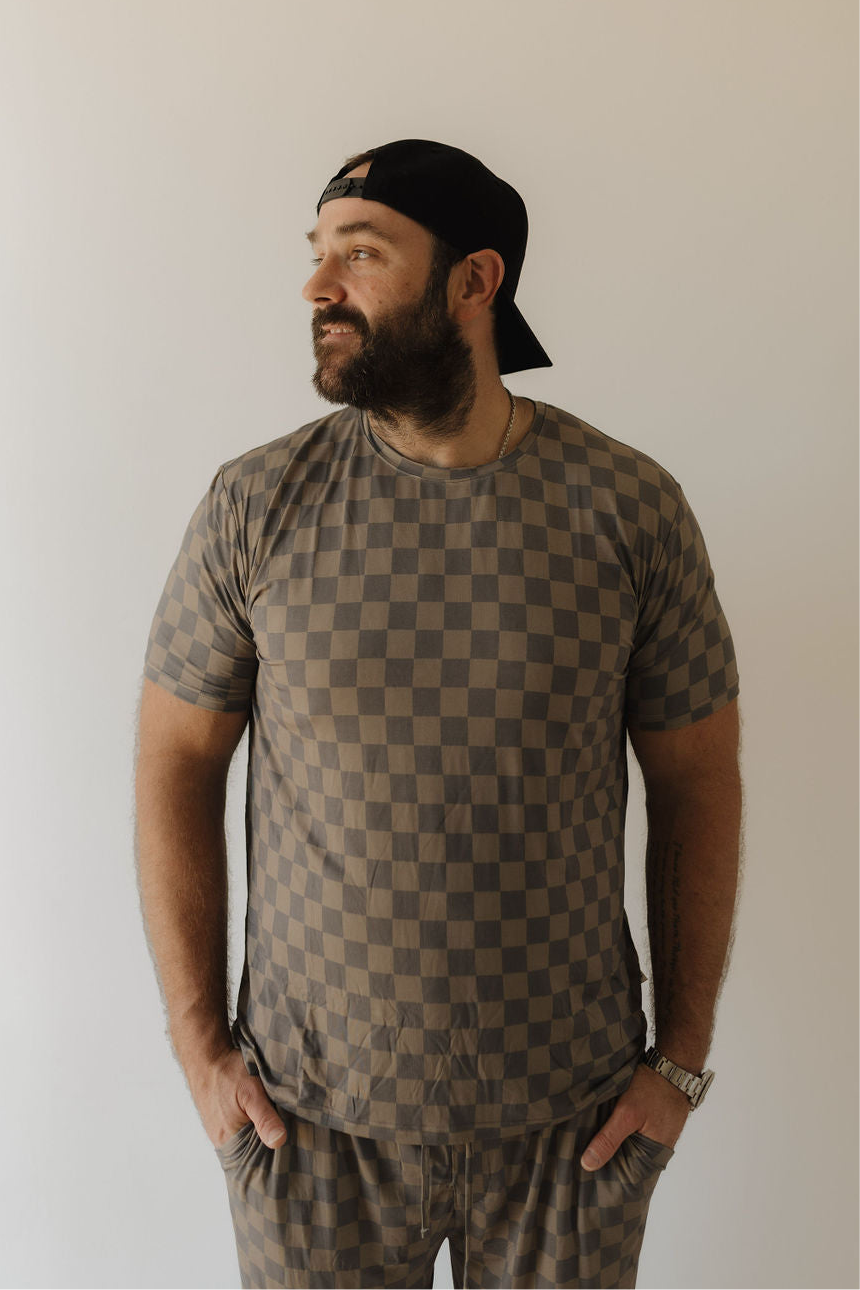 A bearded man in a black backwards cap models forever french baby's Men's Bamboo Short Sleeve Pajama in Faded Checkerboard, standing against a plain light background, looking right with his hands in his pockets.