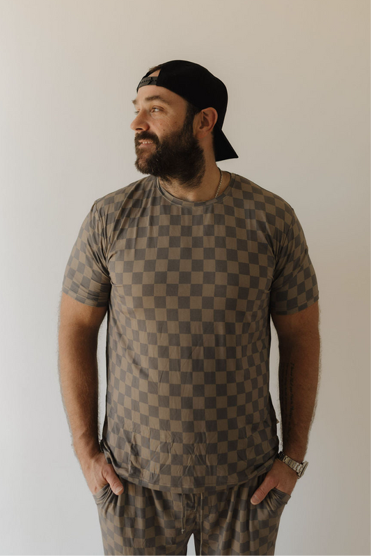 A bearded man in a black backwards cap models forever french baby's Men's Bamboo Short Sleeve Pajama in Faded Checkerboard, standing against a plain light background, looking right with his hands in his pockets.