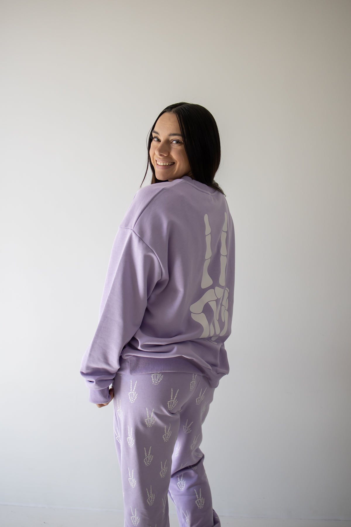 A woman smiles at the camera, standing against a plain light-colored wall in the forever french baby Women’s Sweat Set | Enchanted Skelly—a cozy lavender set adorned with white hand symbols, making it the perfect choice for a spooky season outfit.