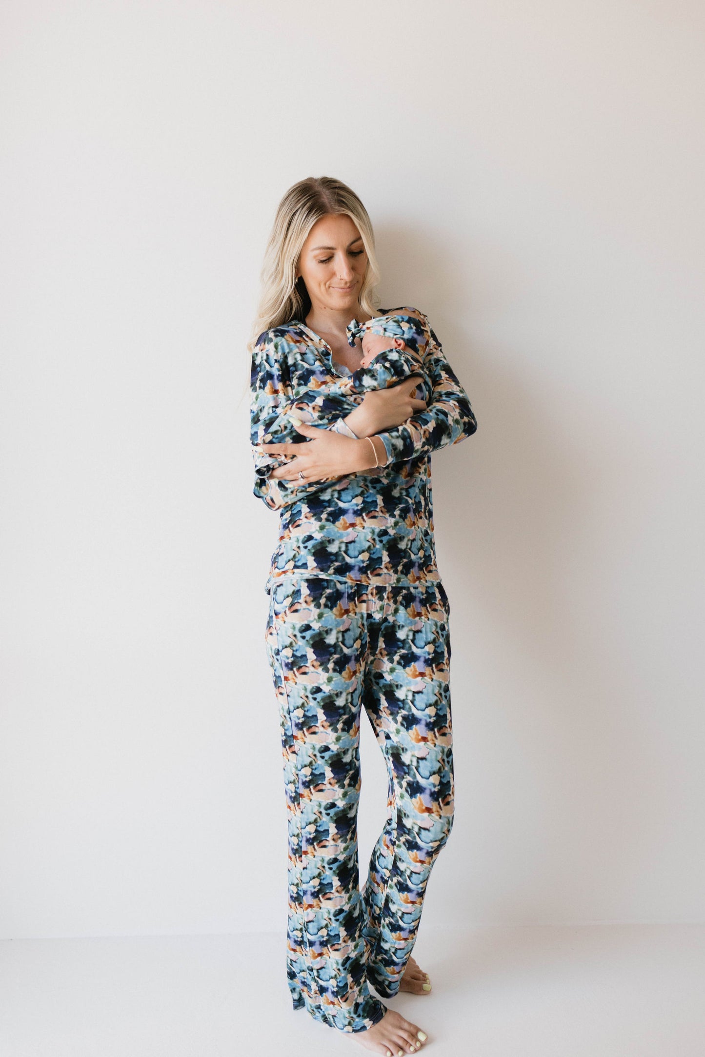 A woman stands against a plain white background, holding a baby close to her chest. She is dressed in matching Charli Print bamboo pajamas from the brand forever french baby, and she is barefoot. The woman gazes down affectionately at the baby in her arms.