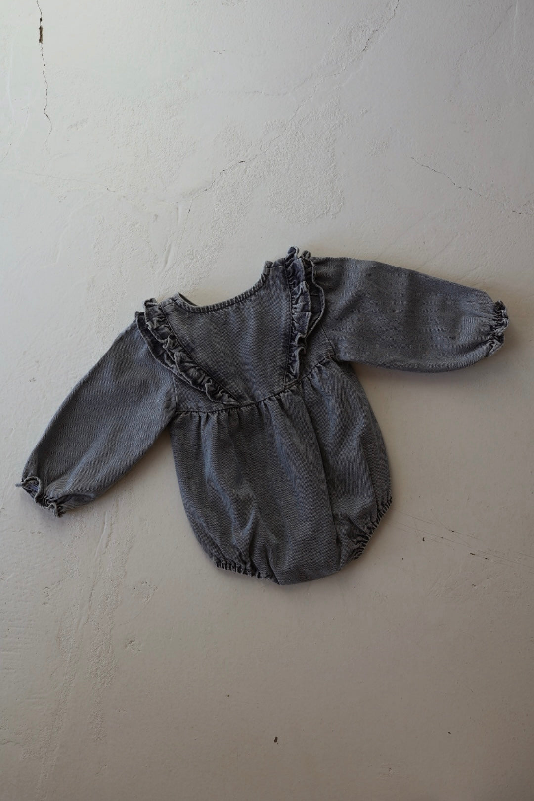The forever french baby Denim Romper | Ruffle features long sleeves, chest and shoulder ruffles, gathered cuffs, and elastic leg openings for a tunic-style look, shown laid flat on a light textured surface.