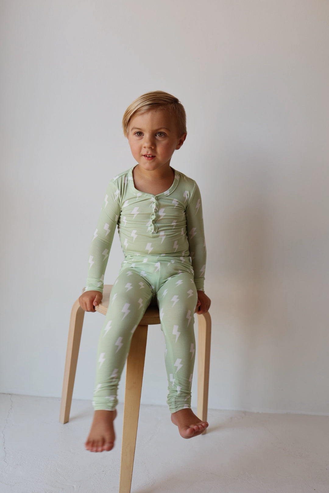 A young blond child sits barefoot on a wooden stool, dressed in forever french baby's Bamboo Two Piece Pajamas | Flash—light green pajamas with white lightning bolt patterns—against a plain, light background.