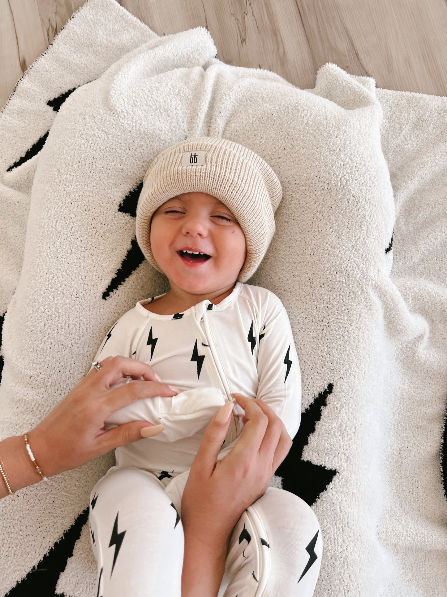 A young child lies on a fuzzy white blanket with black lightning bolt designs, wearing a beige knit hat and Forever French Baby Bamboo Zip Pajamas in White & Black Lightning Bolt pattern. The child is smiling joyfully as someone, partially visible, tickles their tummy.