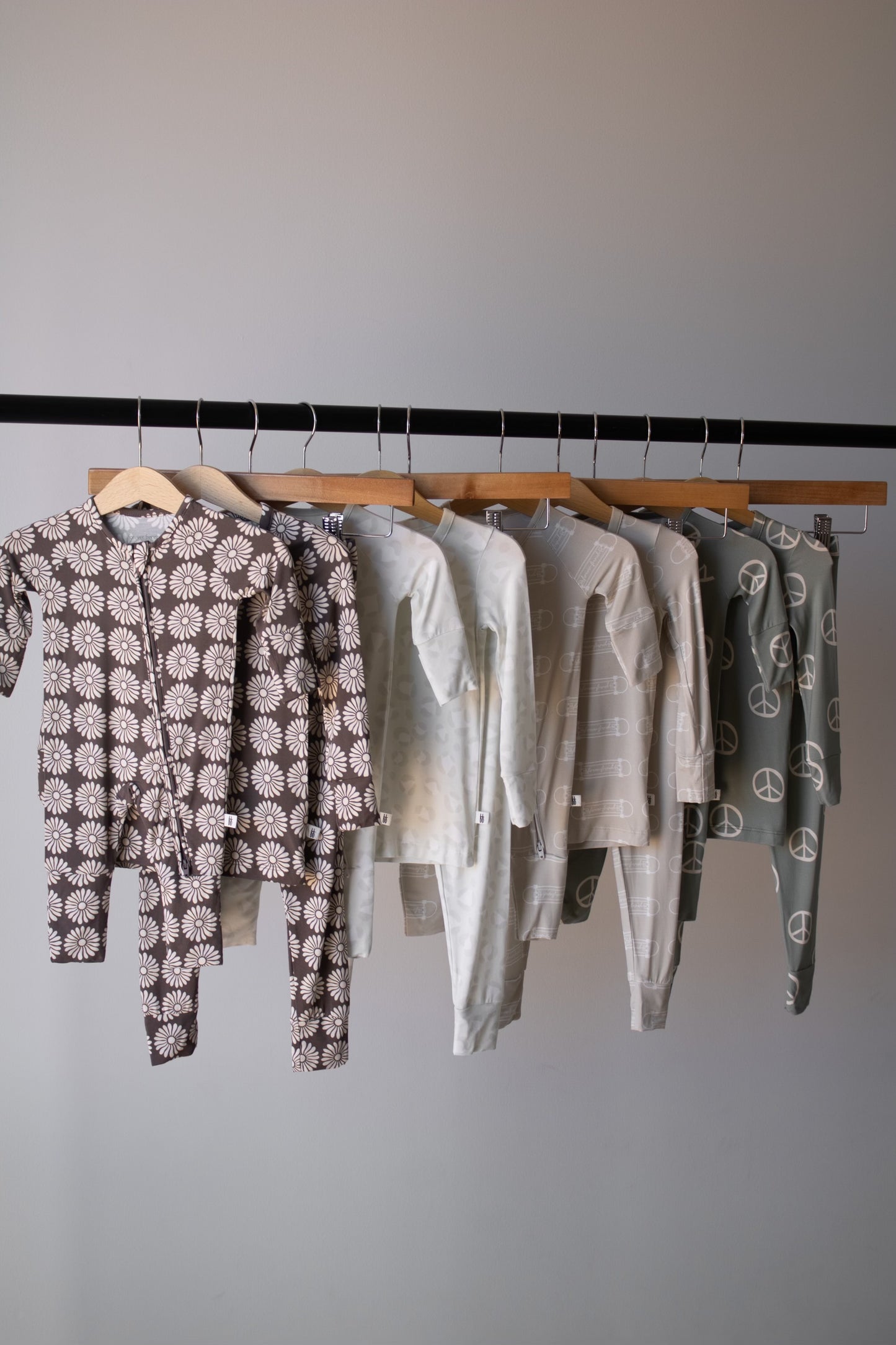 Six sets of forever french baby Bamboo Two Piece Pajamas | Sk8er Boy in neutral tones hang on a rack, each featuring breathable fabric and playful patterns like daisies, stars, stripes, and peace signs.