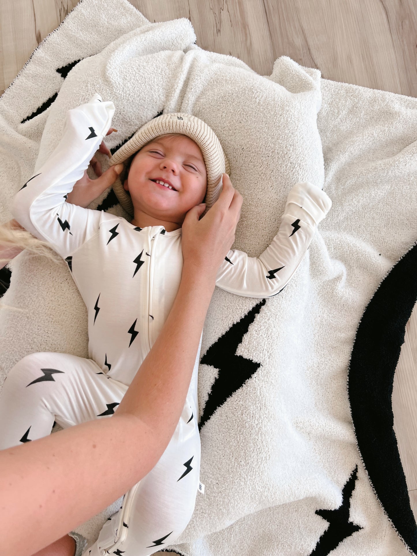 A young child is lying down on a soft, white blanket with black lightning bolt designs. The child, dressed in Forever French Baby’s Bamboo Zip Pajamas in the White & Black Lightning Bolt pattern, smiles with eyes closed while an adult adjusts a beige knit hat on their head. The background is a light wooden floor.