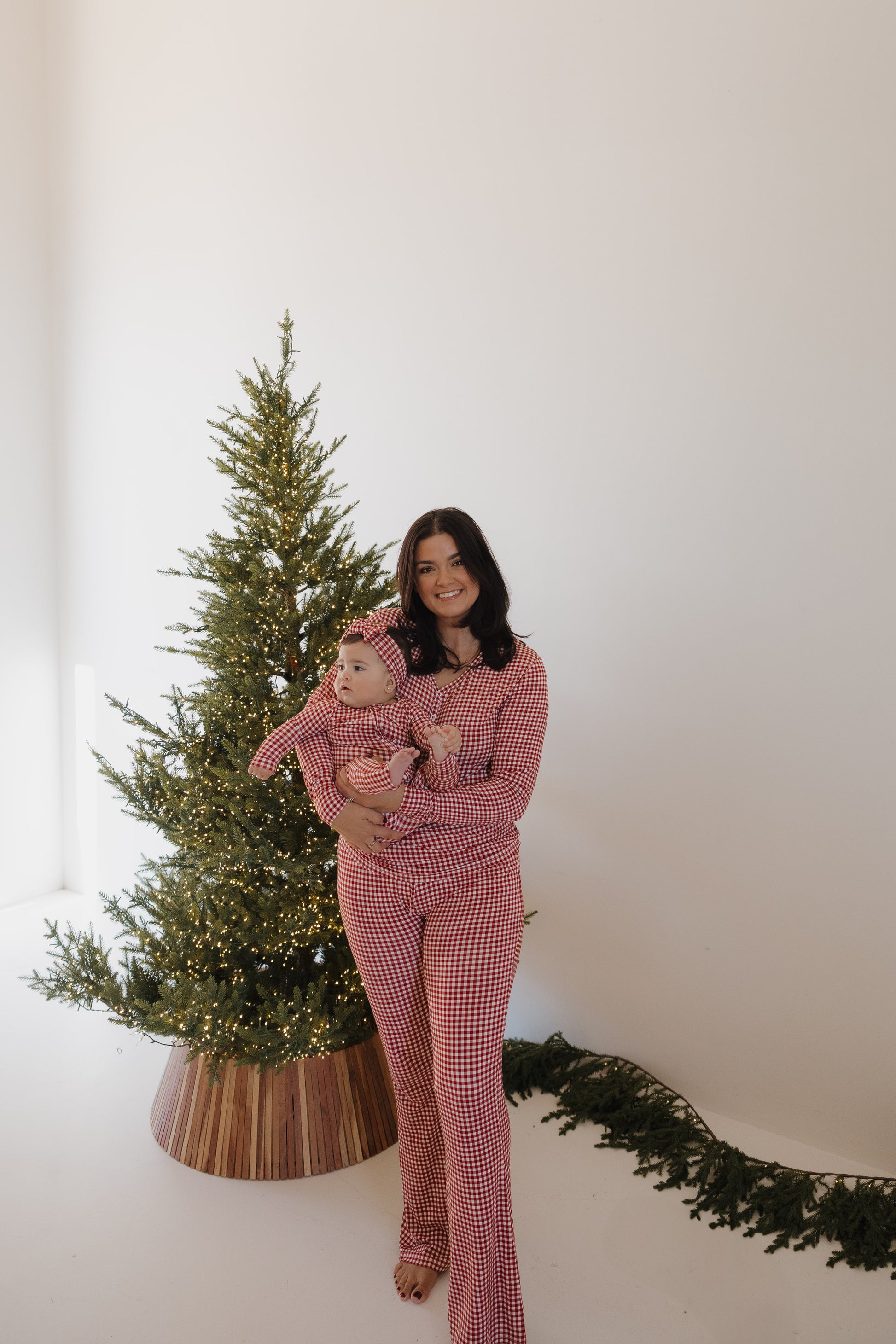A woman holds a baby beside a Christmas tree, both wearing forever french baby's Women's Bamboo Pajamas | Red Gingham, known for hypo-allergenic bamboo fabric. The scene features a minimal white wall and greenery on the floor.