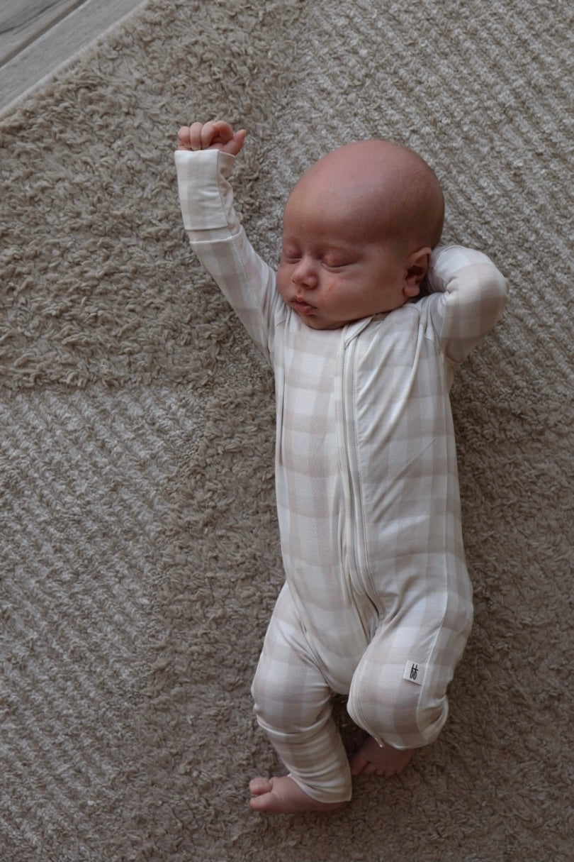 A baby in forever french baby Bamboo Zip Pajamas | Gingham—breathable, hypoallergenic beige and white plaid sleepwear—sleeps on a textured beige carpet, one arm raised and the other bent, lying on its back.