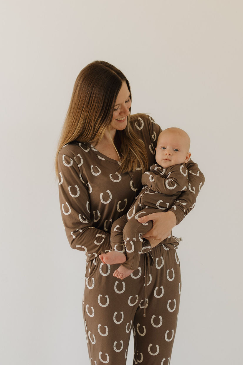 A woman holds a baby as they both wear matching "Giddy Up" brown bamboo pajamas with white horseshoe patterns from forever french baby, standing against a plain light background.