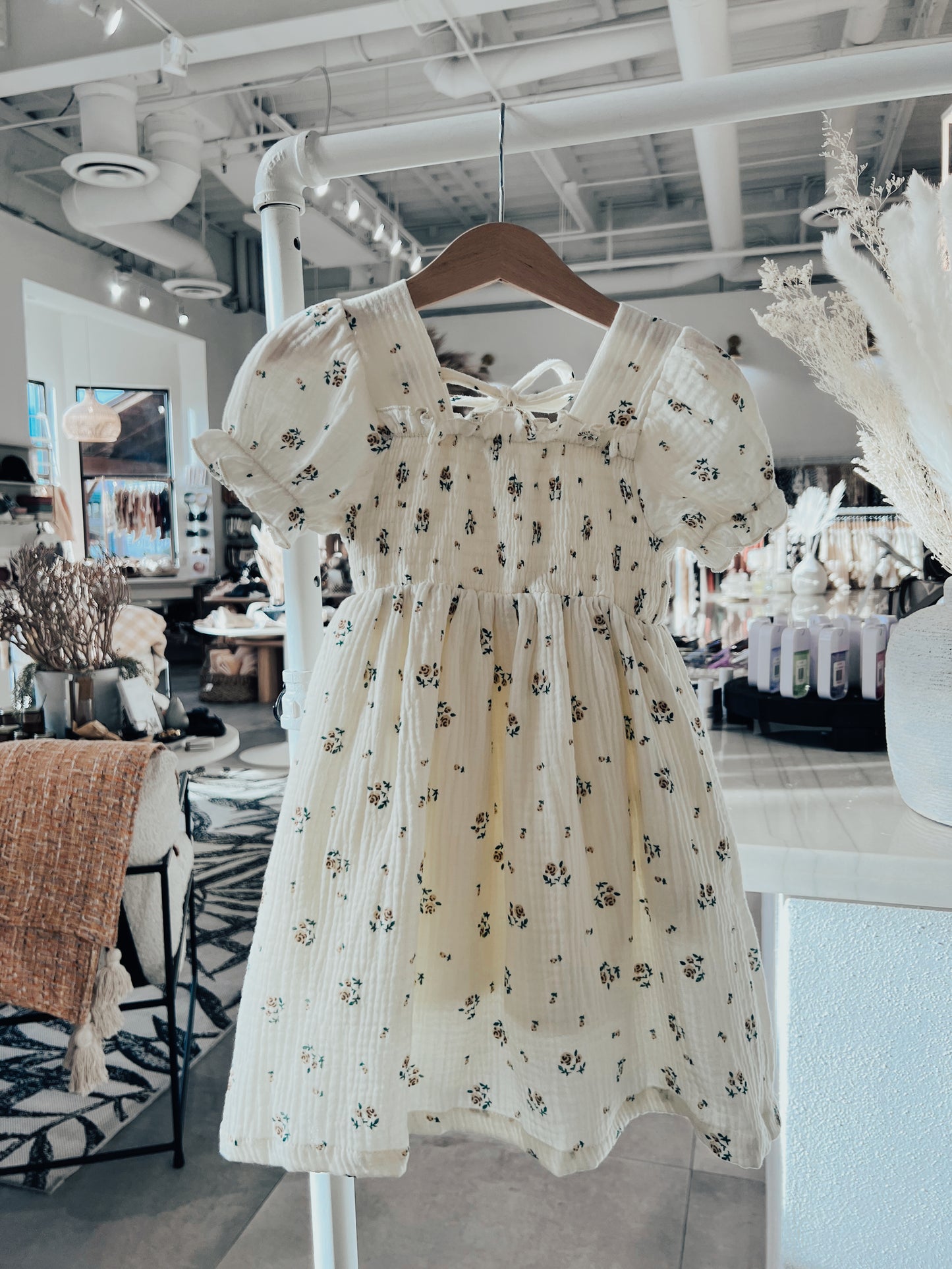 A light yellow dress with a floral print and puffed short sleeves, from the "Child & Women's Muslin Dress | Garden Days" collection by forever french baby, hangs on a wooden hanger in a brightly lit boutique. This charming dress, featuring a ruched bodice, is part of the matching mom and daughter dresses collection and is displayed among various home decor and clothing items in the background.