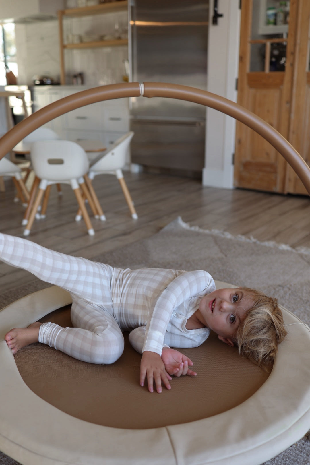 A young child in forever french baby Bamboo Two Piece Pajamas | Gingham lies on their side on a round, padded play mat with a wooden arch in a modern kitchen and dining area.