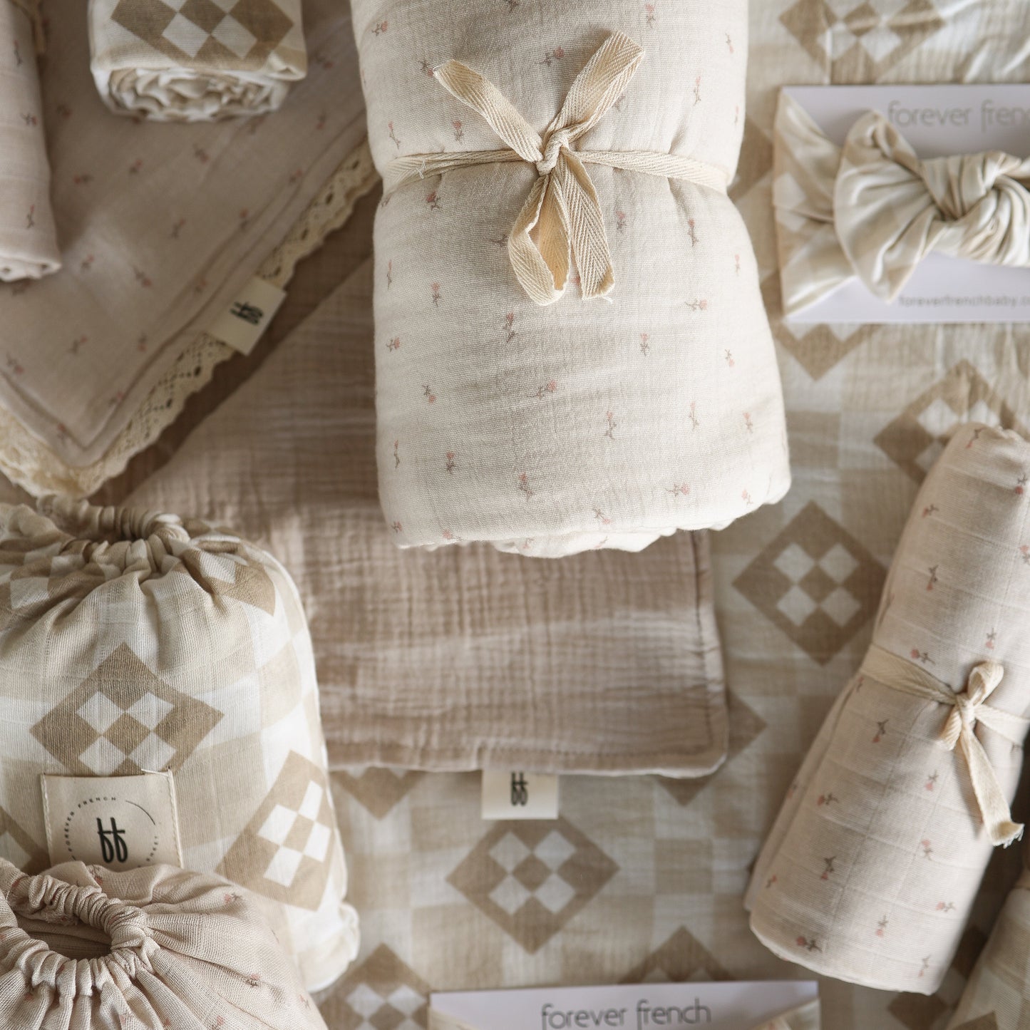 A selection of soft, beige Muslin Quilt | Lucy blankets from forever french baby, some rolled and tied with ribbon, displayed on a quilt-patterned fabric background with "forever french" labels.