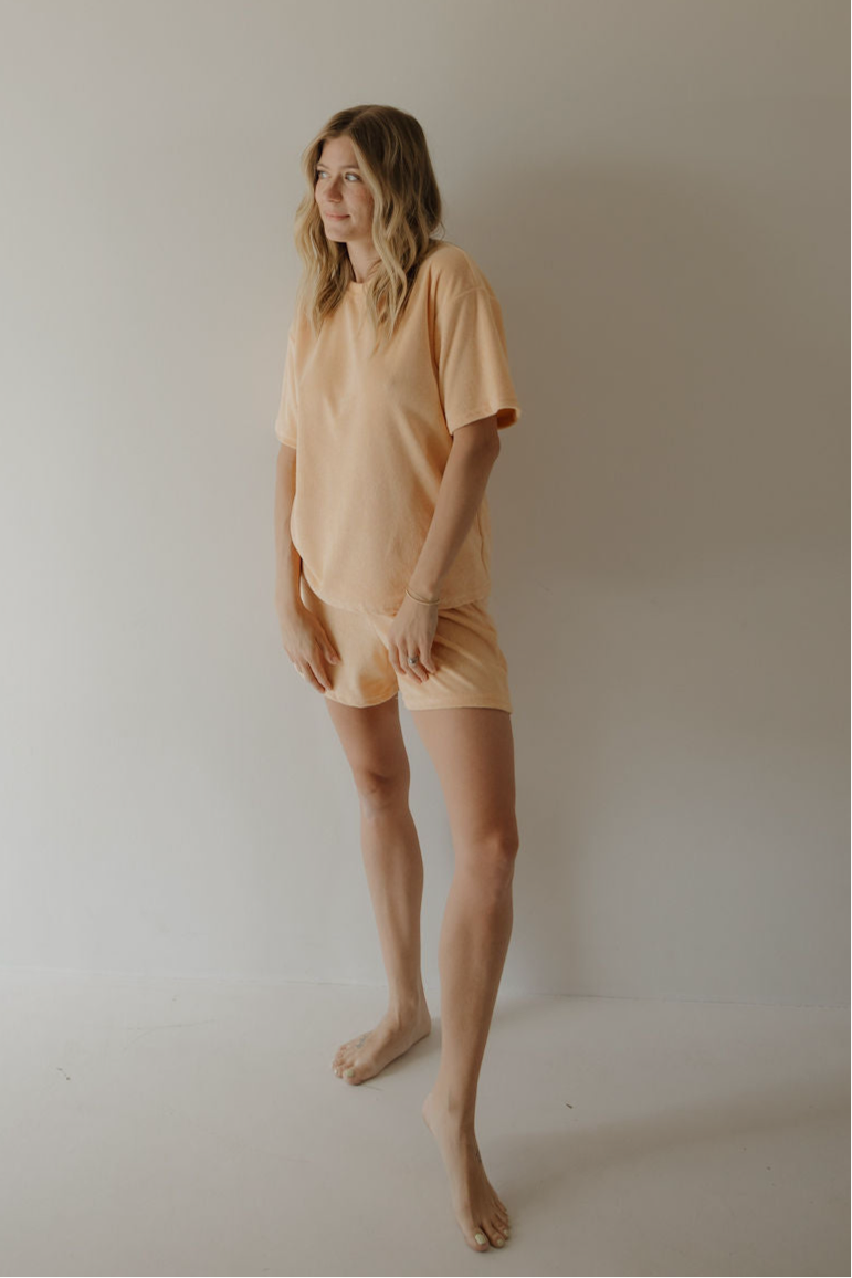 A woman with wavy blonde hair, barefoot against a light wall, wears the forever french baby Women's Terry Short Set in Sherbert, looking slightly to the side and smiling.