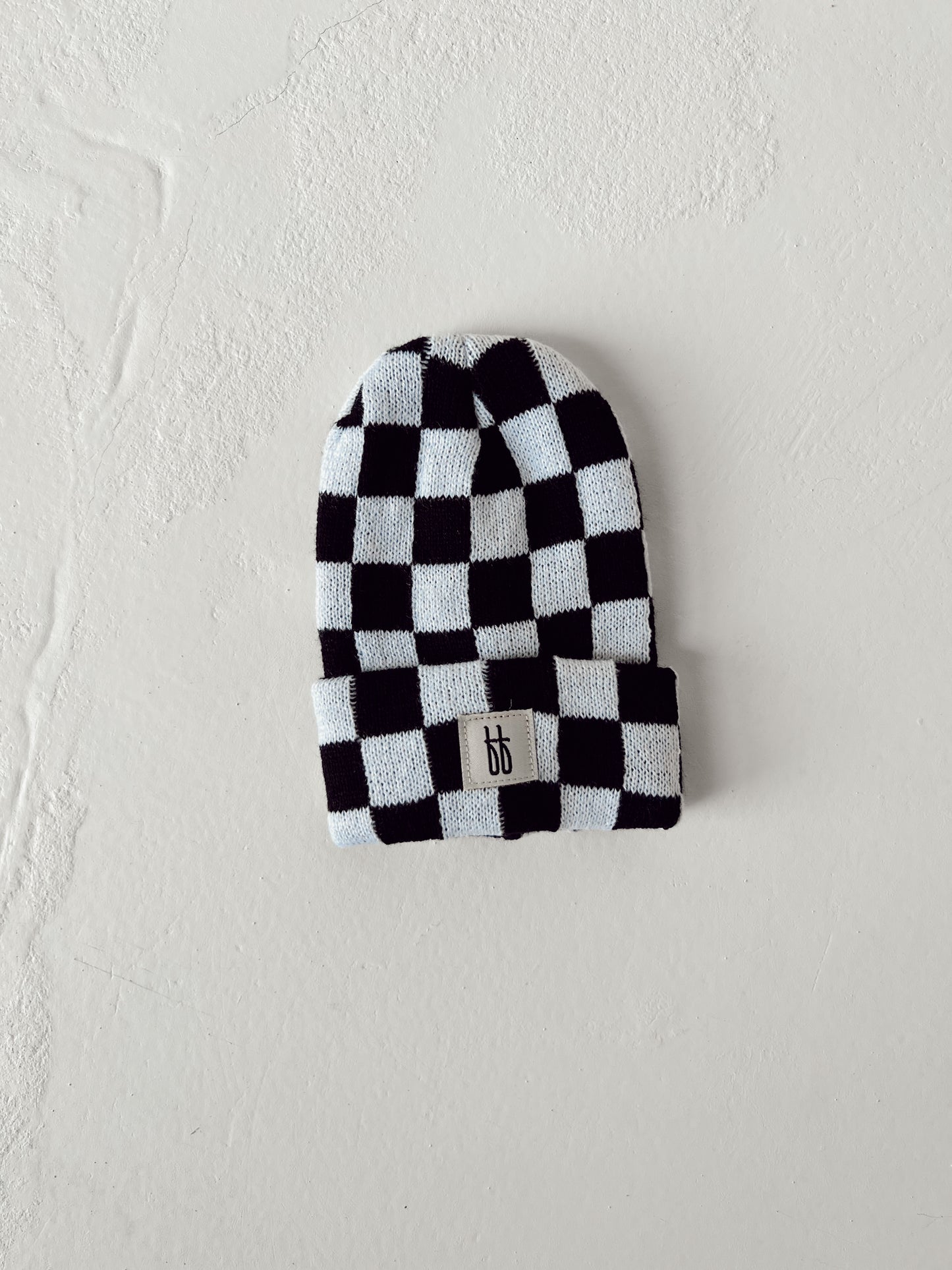 A black and white checkerboard-patterned ff Knit Beanie from forever french baby, featuring a small rectangular logo tag with stylized initials "ff" at the front. Made from breathable yarn, the hat is laid flat against a textured off-white background.