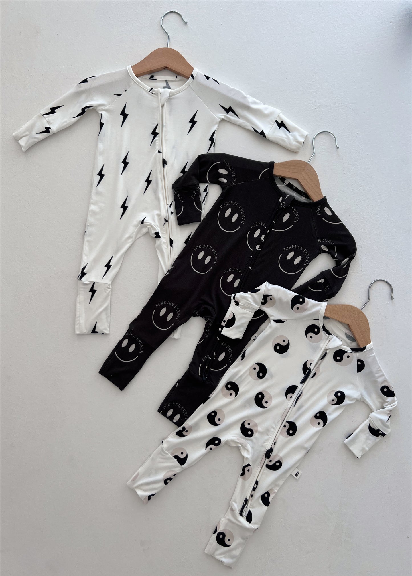 Three baby onesies on hangers are displayed against a plain background. The first is white with black lightning bolts, the second is black with smiling faces and the text "Whatever," and the third is white featuring a stylish Black & White Yin Yang design. This adorable Baby and Toddler clothing from "forever french baby" showcases comfort and style for your little one, especially with the Bamboo Zip Pajamas.