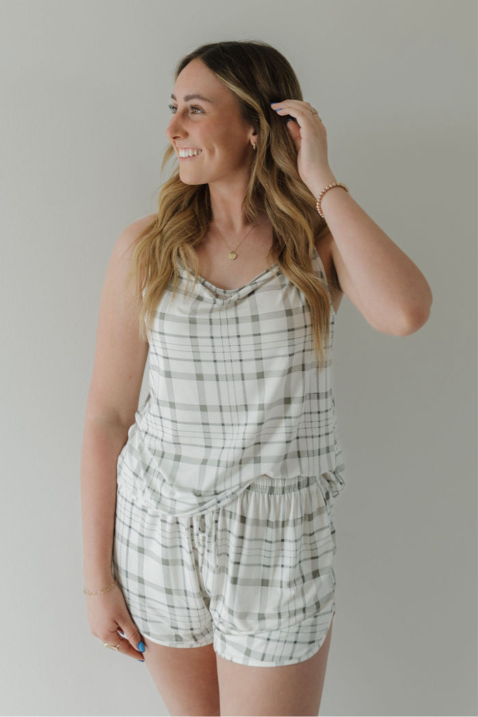A woman stands against a plain light wall, smiling to the side and touching her hair, wearing the forever french baby Cami Women's Bamboo Set in Picnic Plaid—a sleeveless, breathable plaid tank top with matching shorts.