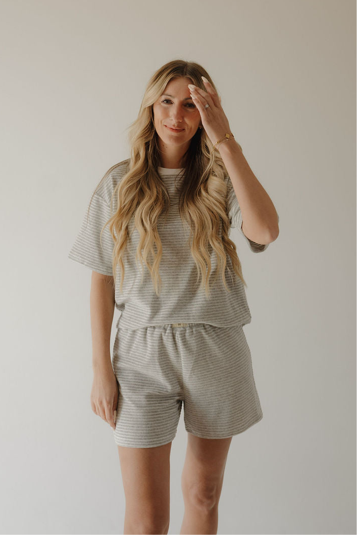 A woman with long, wavy blonde hair wears the forever french baby Women's Terry Short Set in grey stripe, standing against a plain background and smiling gently while raising one hand near her face.