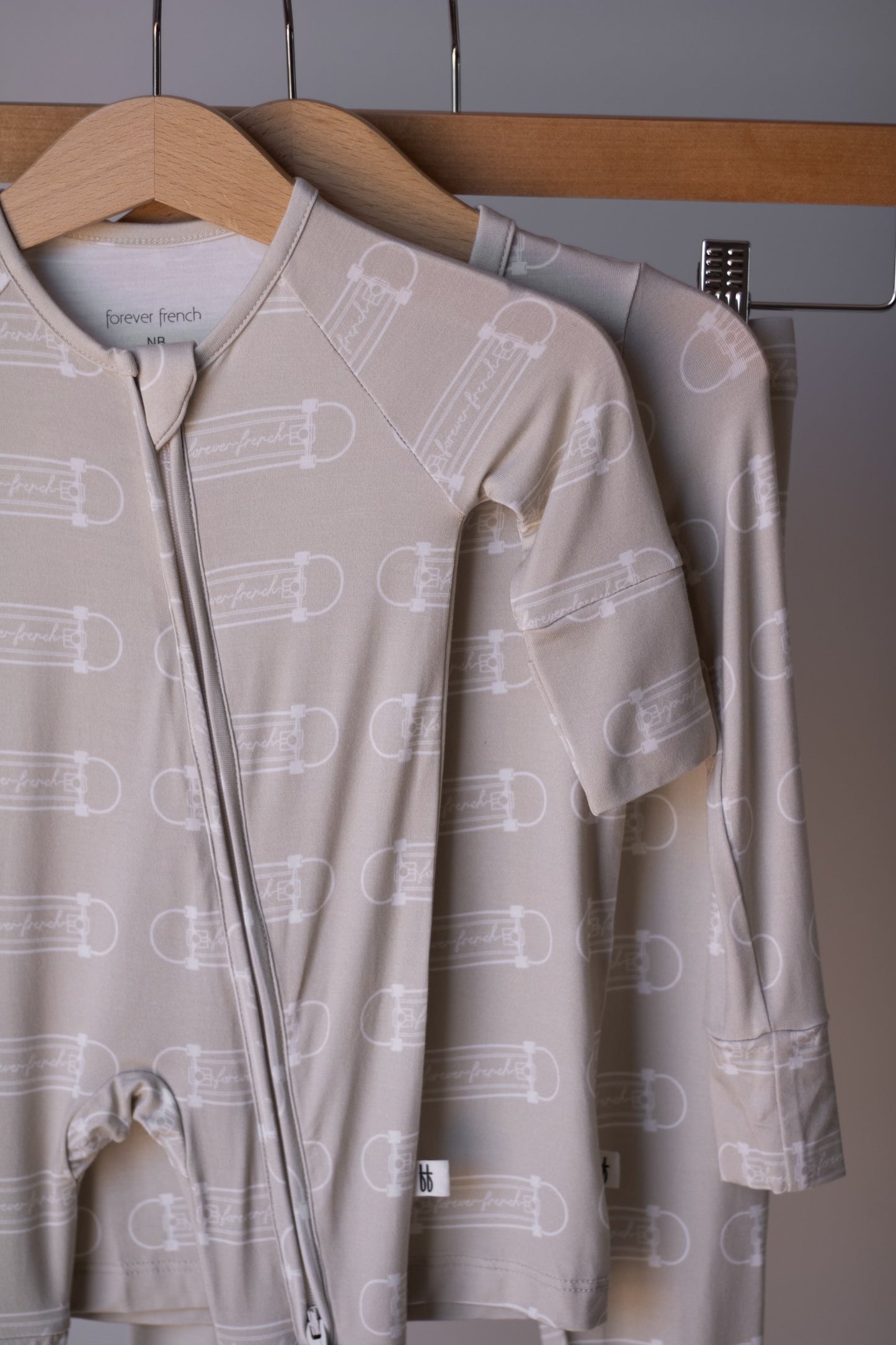 Two beige "Bamboo Two Piece Pajamas | Sk8er Boy" by forever french baby hang on wooden hangers from a metal rod. The front pajama is made from breathable fabric, features a zipper, and has a "forever french baby" tag.