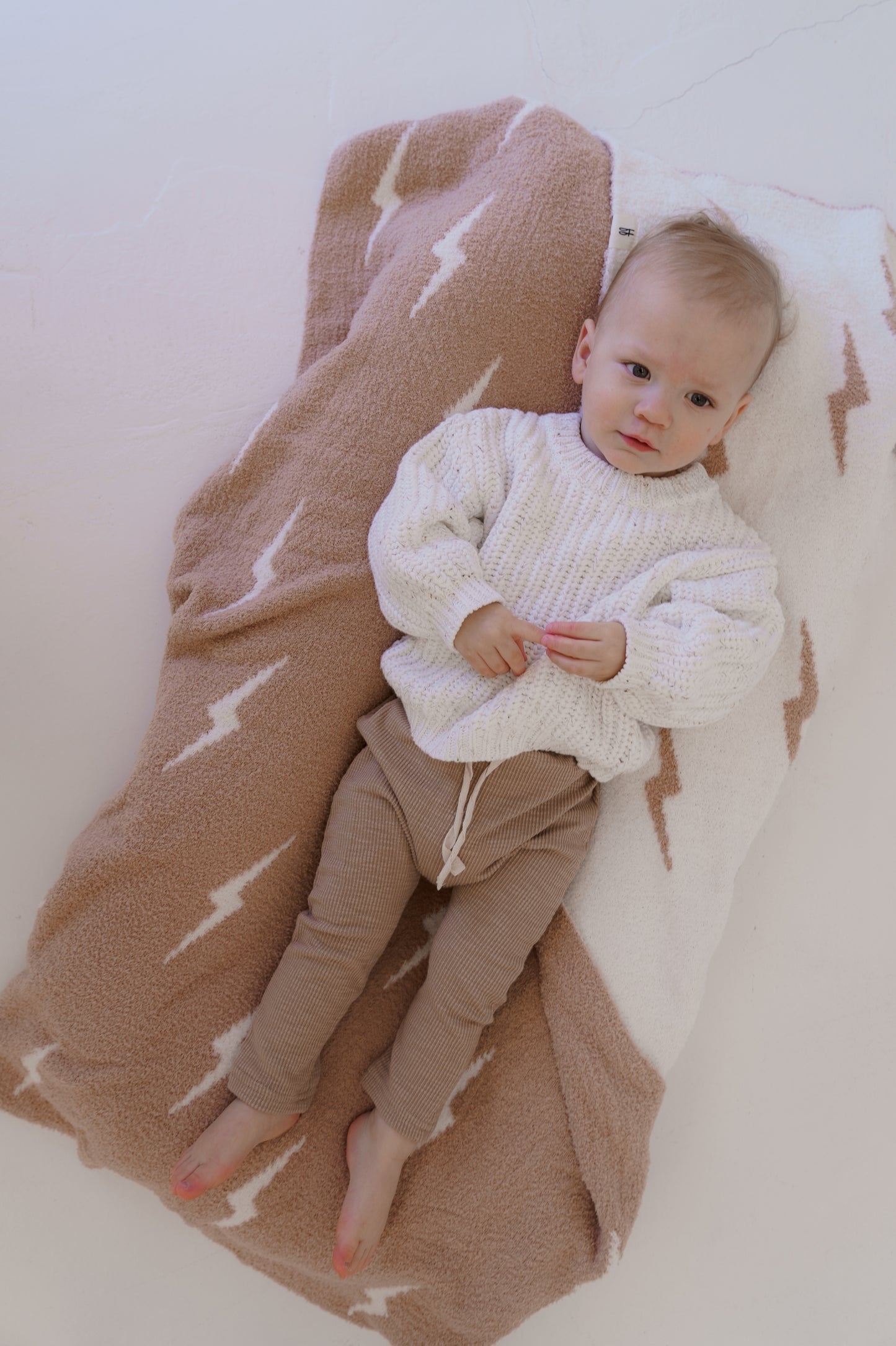 A baby rests on a soft brown and white zigzag-patterned blanket, wearing a cream knit sweater from forever french baby's Cloud Confetti line and brown pants, with bare feet. The background is smooth and white.