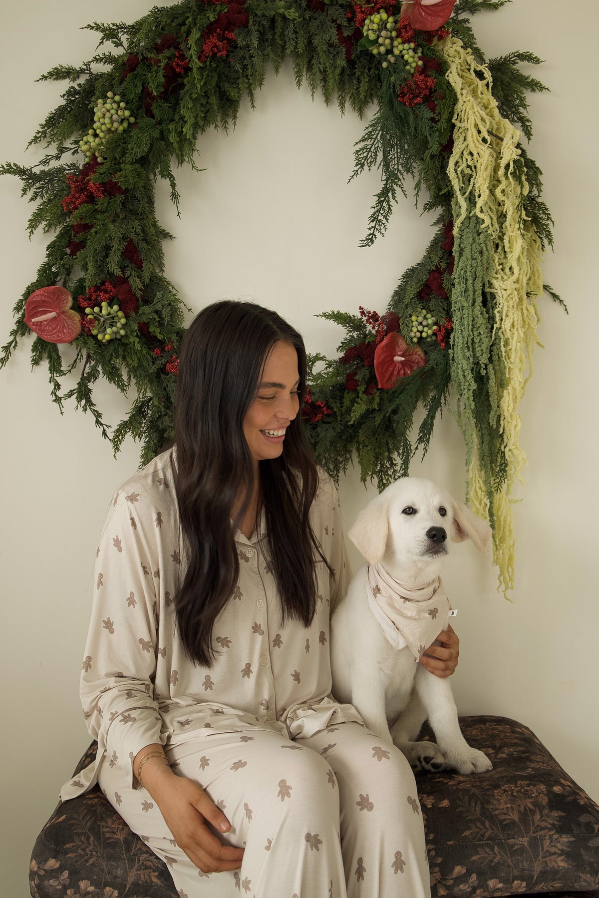 A smiling woman in ginger-cookie-patterned pajamas sits next to a white puppy wearing a Bamboo Bandana | Gingersnap in front of a holiday wreath.