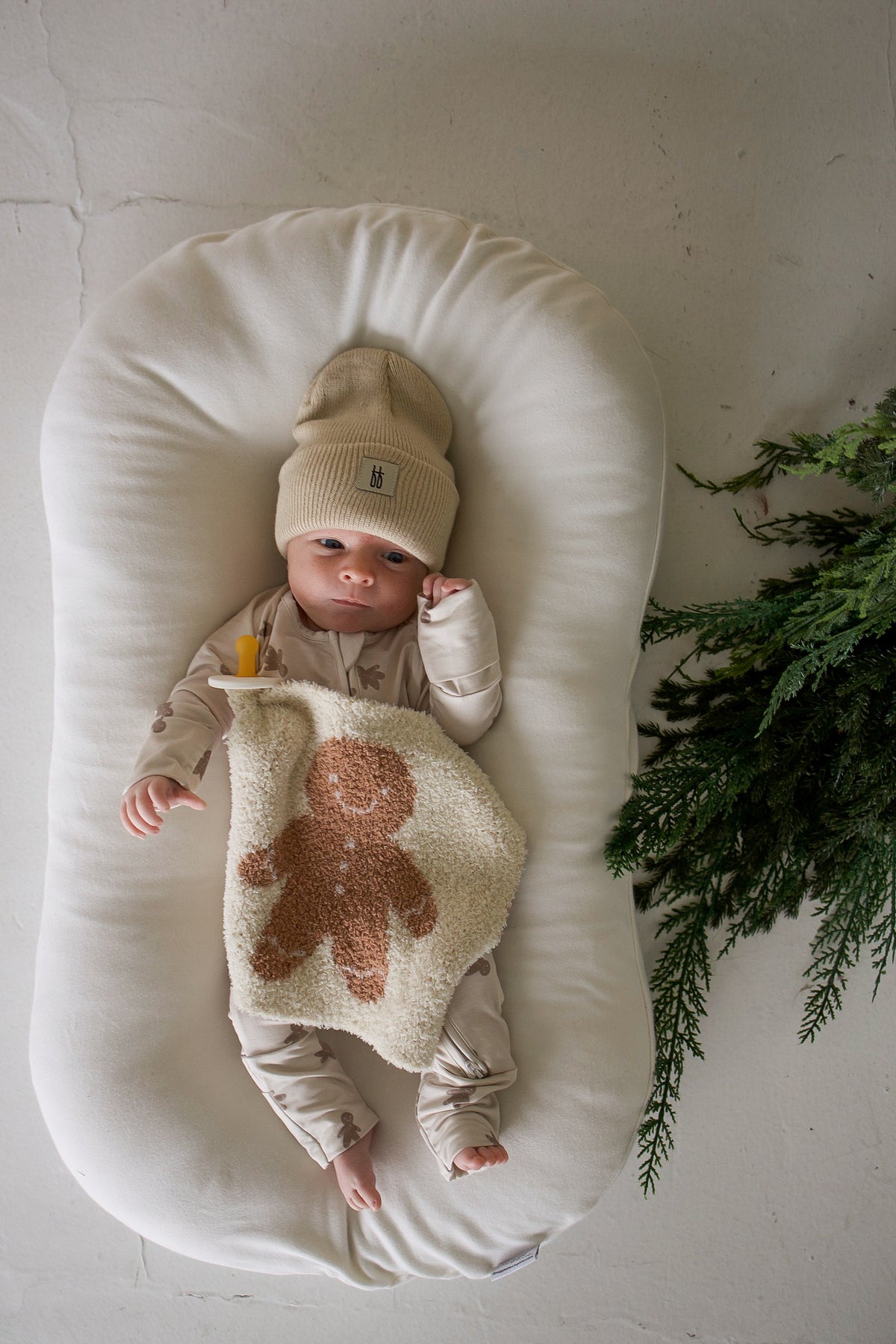 A baby in a beige beanie and matching onesie lies in a white lounger, holding a *PRE-ORDER* Lovey | Gingersnap featuring a textured gingerbread man.