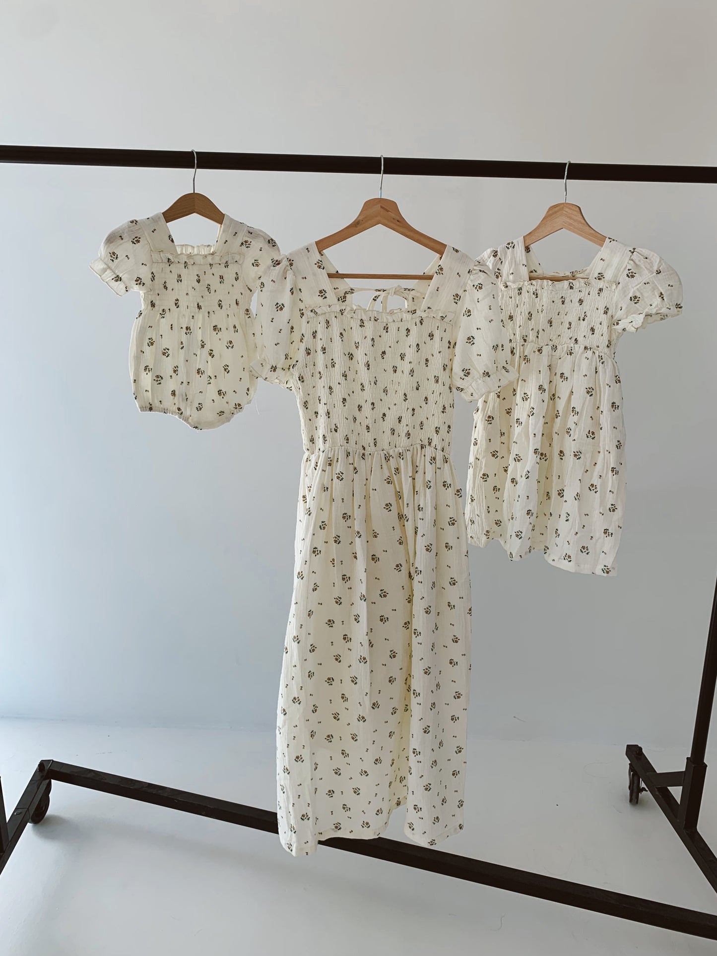 Three light-colored floral print dresses hang on wooden hangers from a black metal garment rack. One is a baby dress, another is from the Child & Women's Muslin Dress | Garden Days collection by forever french baby, and the third is an adult dress—perfect as matching mom and daughter outfits. The background is a plain light gray wall.