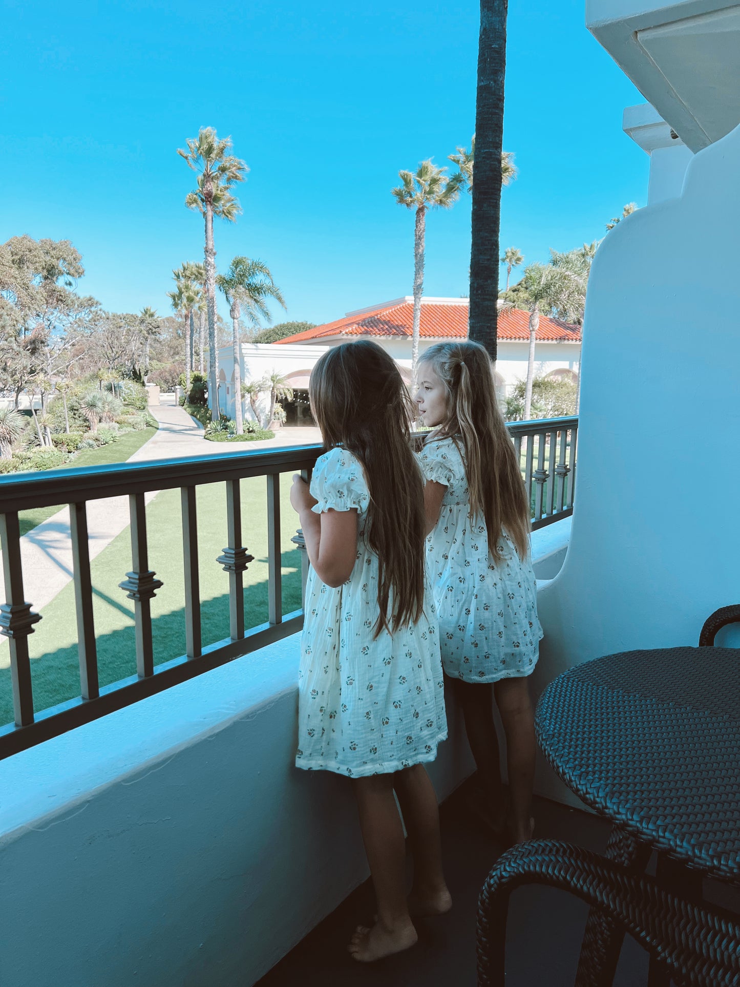 Two young girls, dressed in matching "Garden Days" ivory muslin dresses by forever french baby, stand on a balcony overlooking a sunny landscape with green lawns, palm trees, and Spanish-style buildings with red-tiled roofs. The sky is clear and blue.