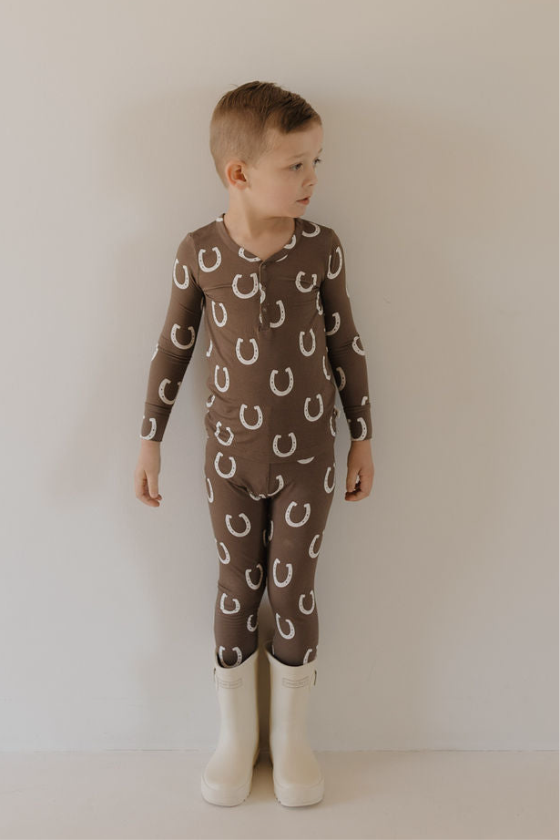 A young child stands against a plain wall, wearing forever french baby’s Bamboo Two Piece Pajamas in the Giddy Up print—brown with white horseshoes—paired with white boots, looking to the side.